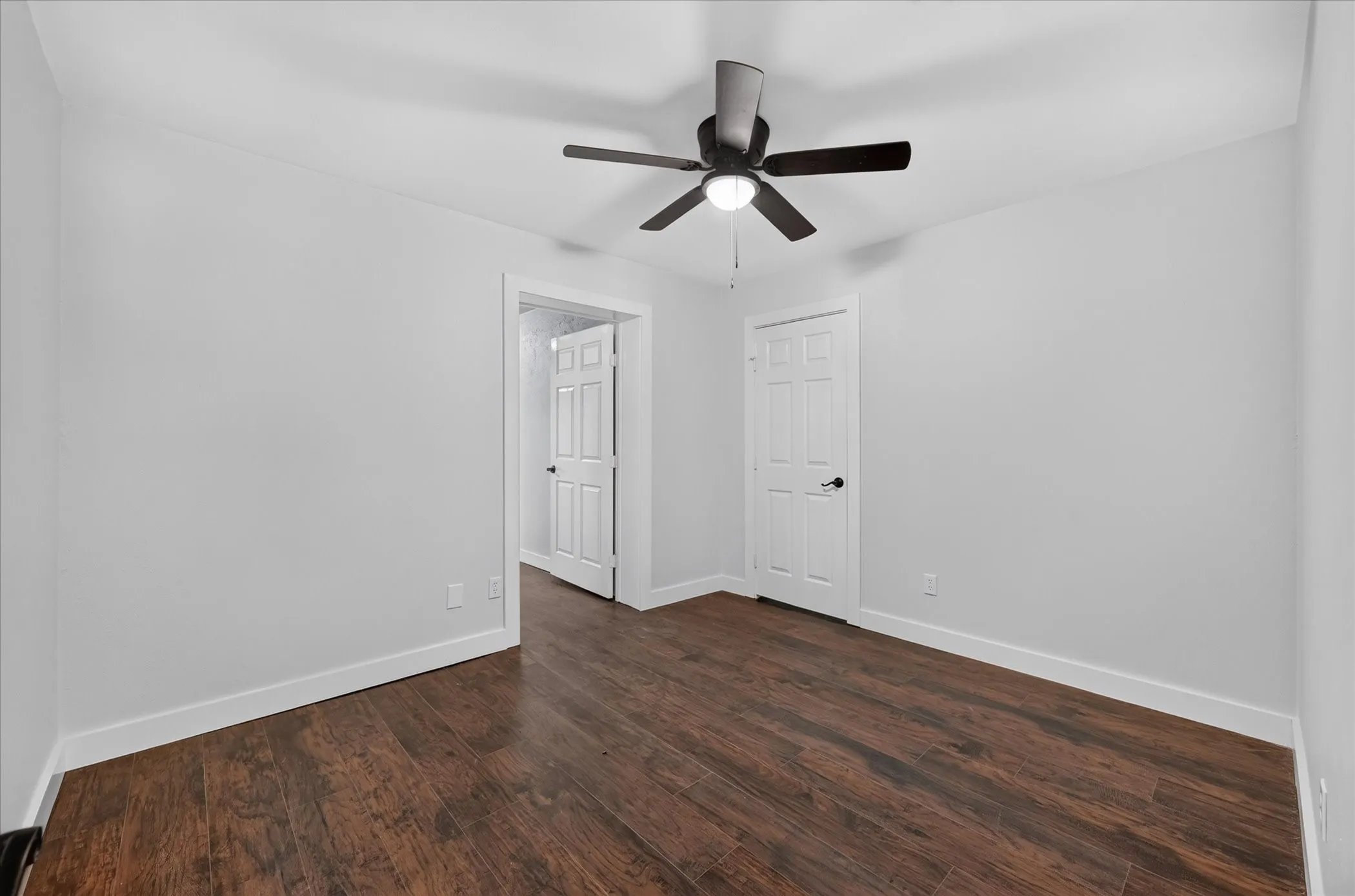 Dark wood-finish flooring with white trim, a ceiling fan with integrated lighting, and a white paneled door with black hardware