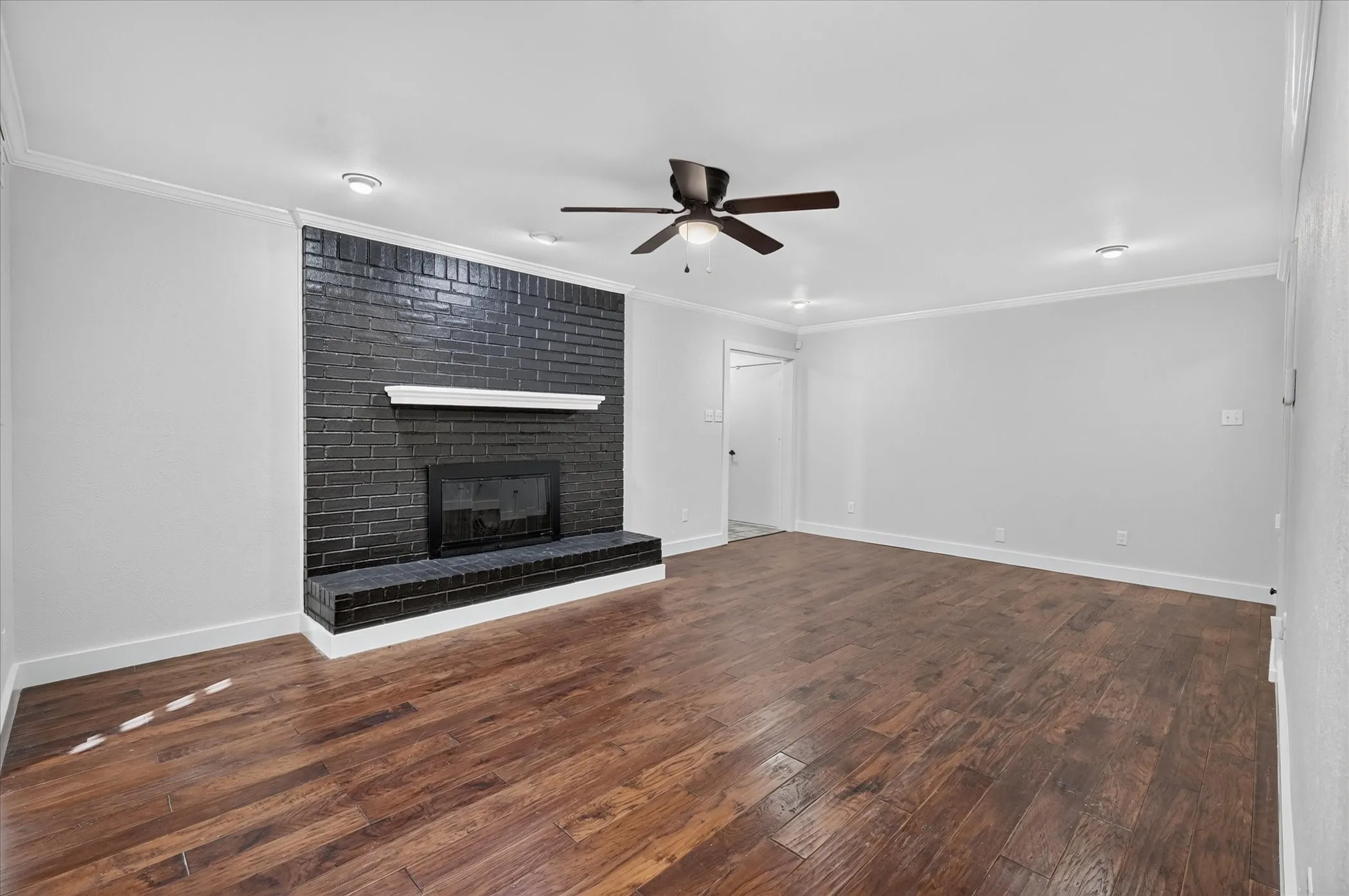 Wood-finish flooring throughout, complemented by a black brick fireplace with a white mantel, a ceiling fan, and recessed lighting