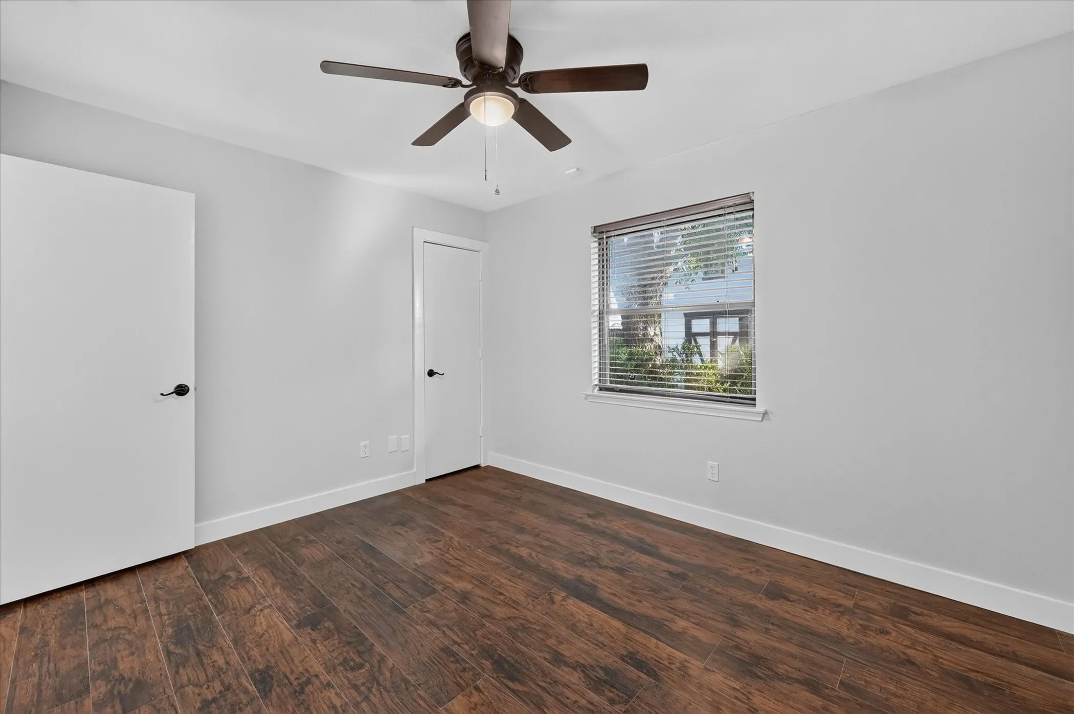 Wood-finish flooring, white walls, two white doors with black hardware, and a ceiling fan with integrated lighting