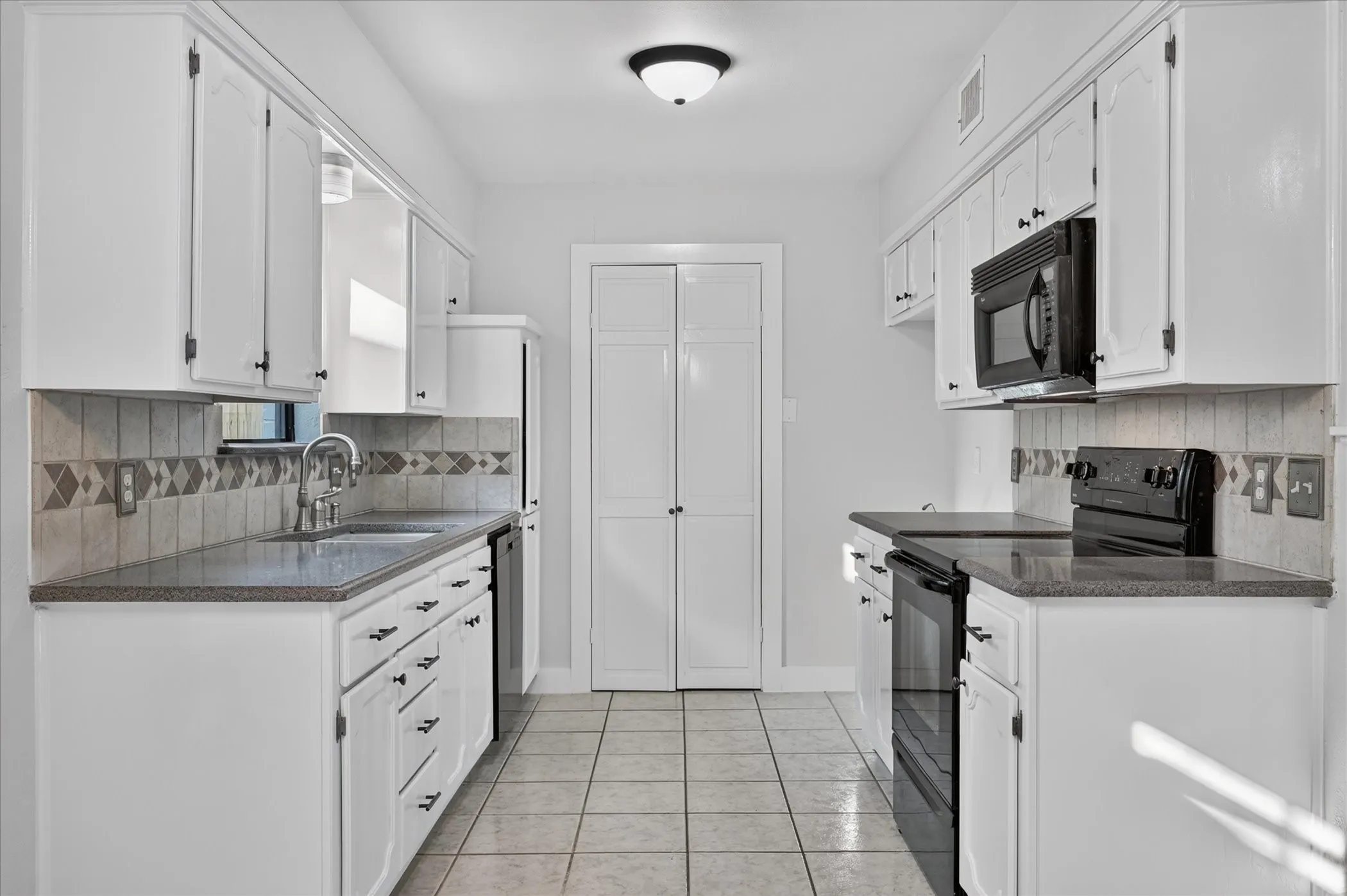 Galley kitchen featuring white cabinetry, dark countertops, tiled backsplash, stainless steel sink, and black appliances