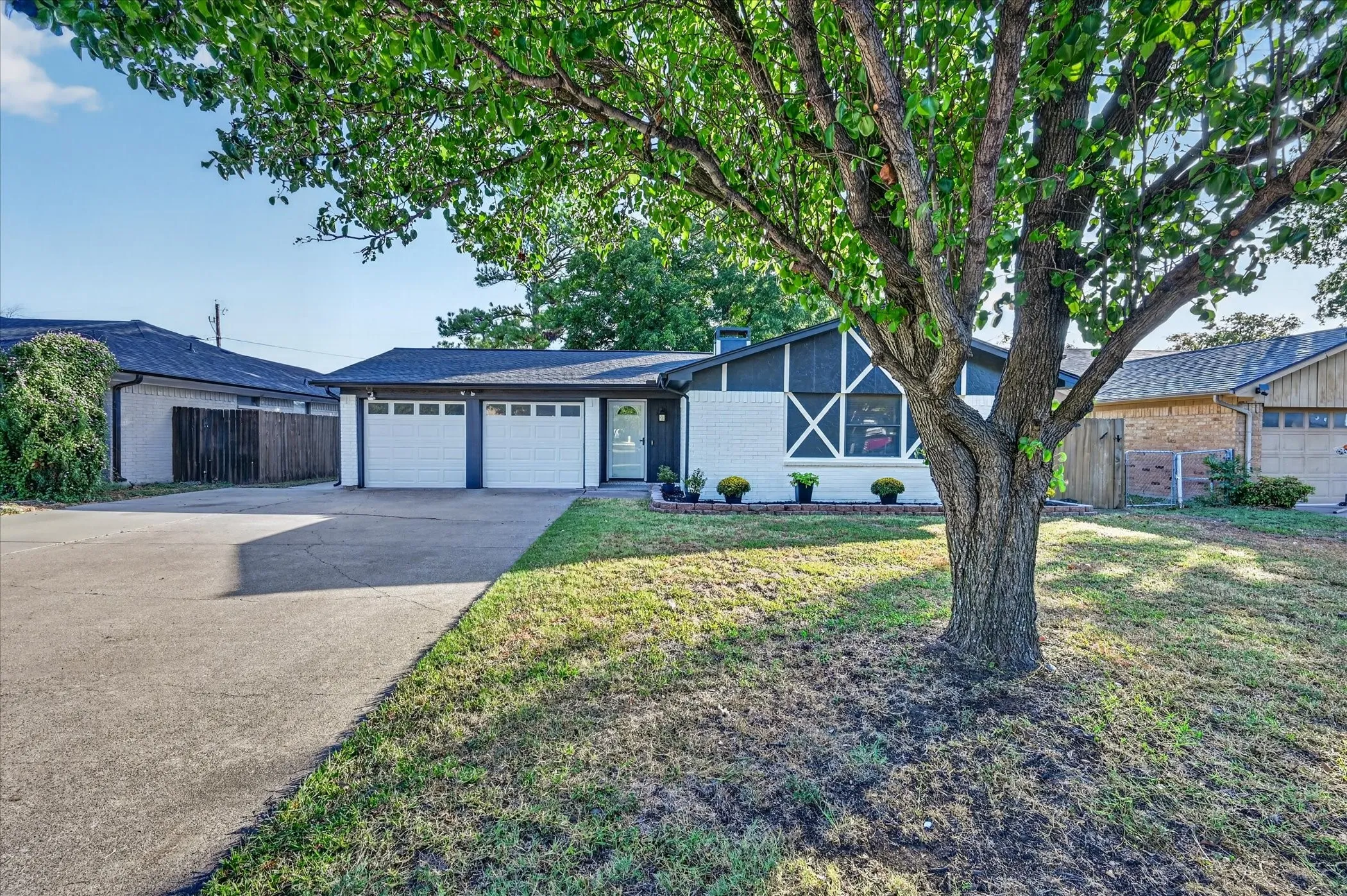 Front exterior featuring a white brick and dark siding facade, a two-car garage with white doors, a concrete driveway, and a mature tree in the foreground