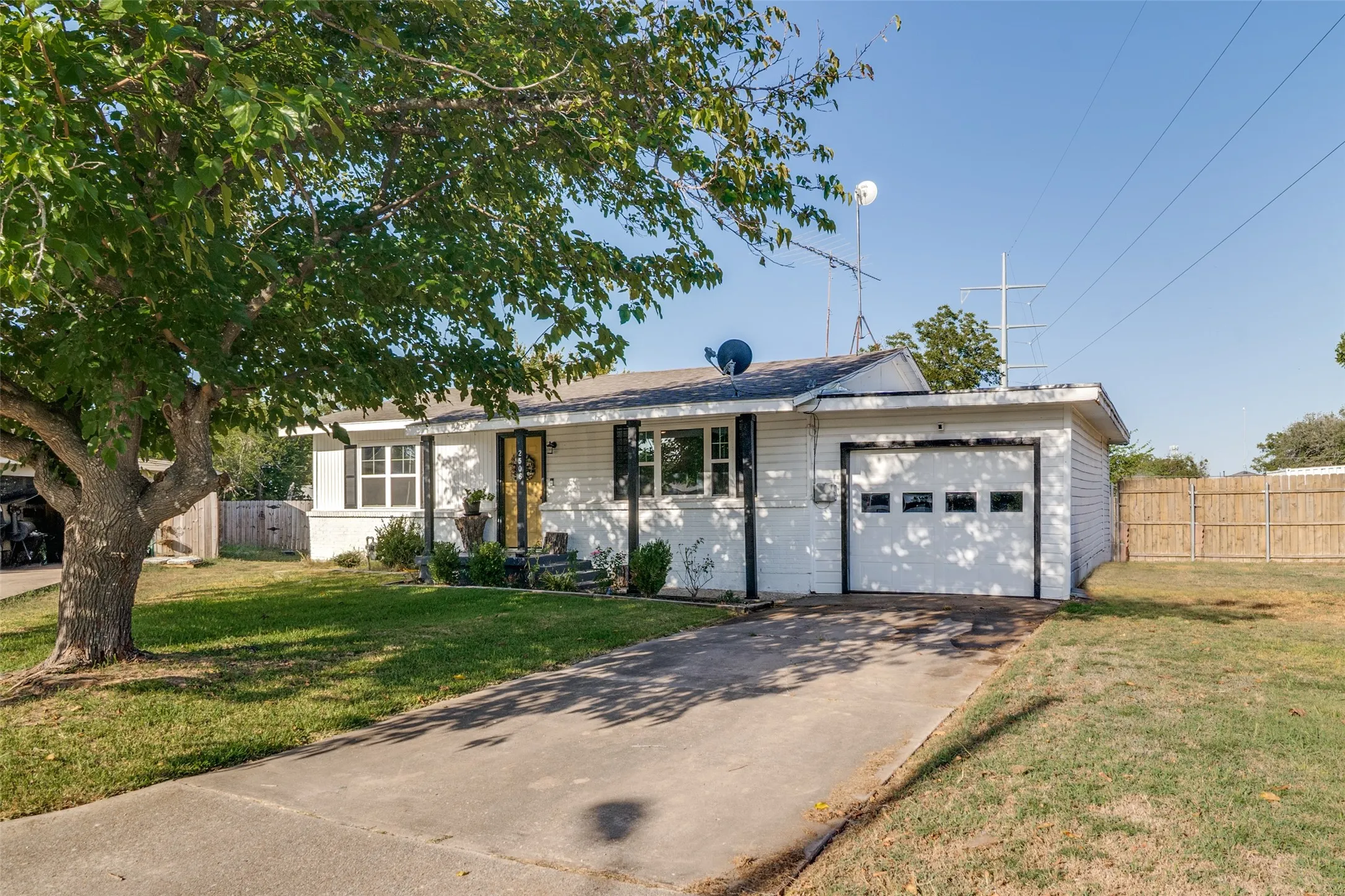 White exterior siding, attached single-car garage, concrete driveway, mature front yard tree, and a wooden privacy fence.