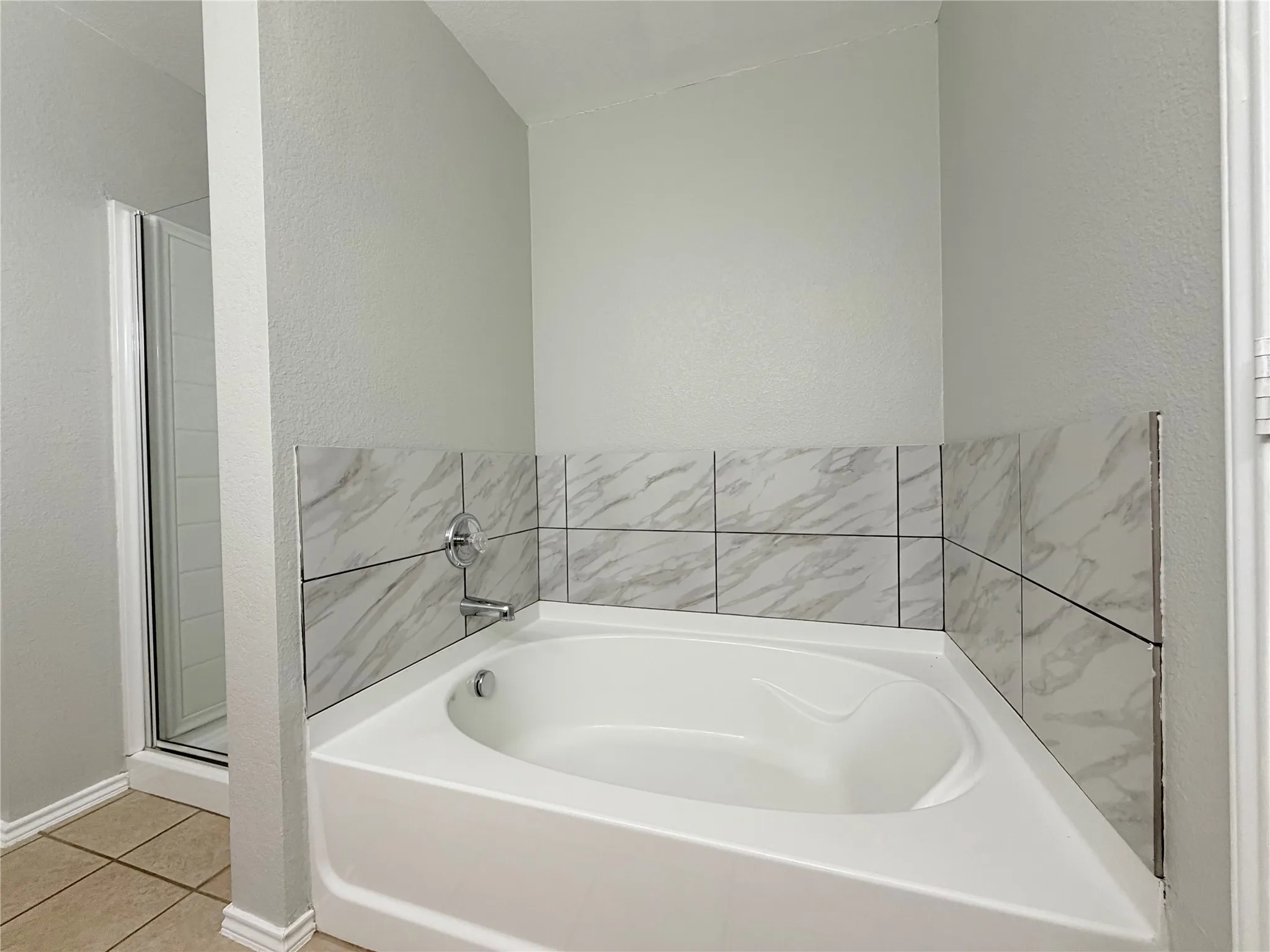 Corner soaking tub featuring a tile surround with marble-look accents and a chrome fixture