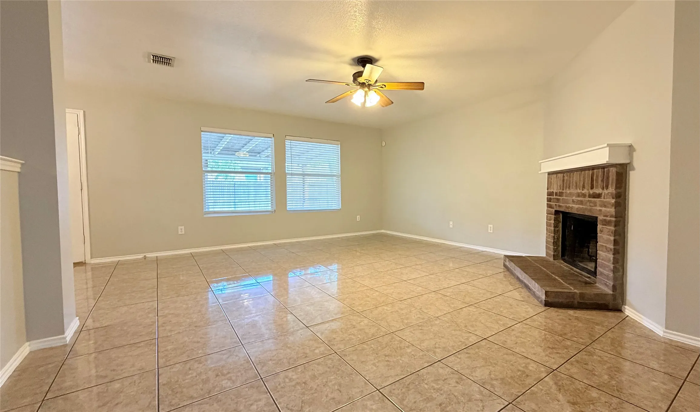 Spacious room featuring tile flooring, a brick-surround fireplace with a mantel, dual windows with blinds, and a ceiling fan