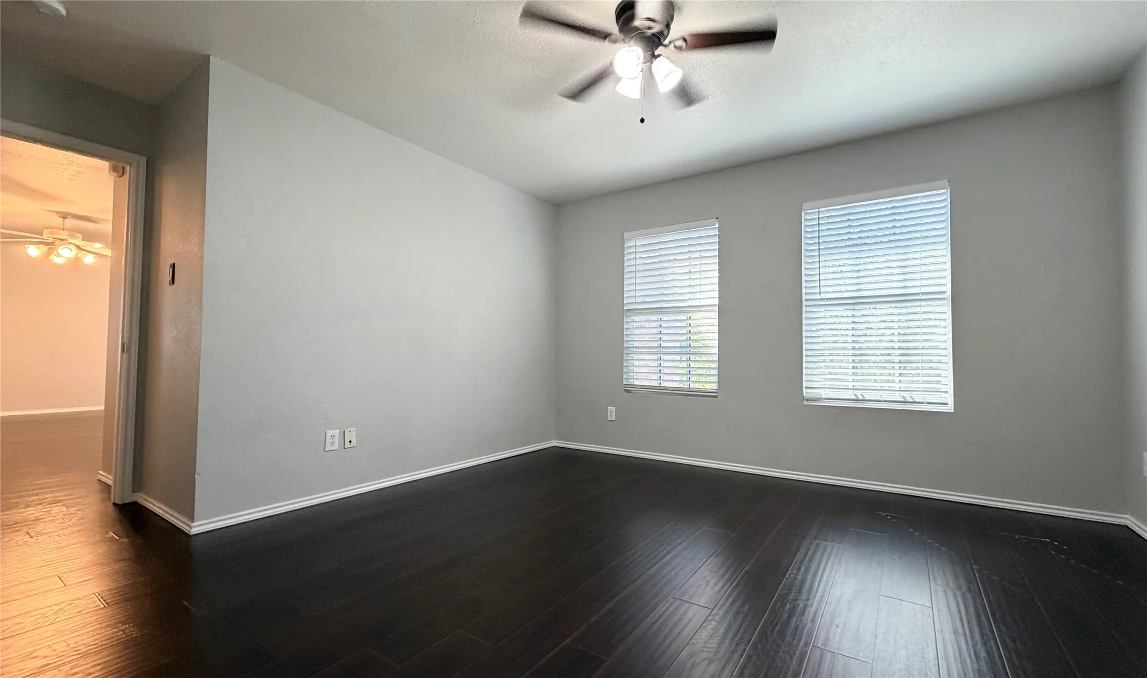 Dark wood-finish flooring, two large windows with blinds, ceiling fan with integrated lighting, and light-toned wall finishes