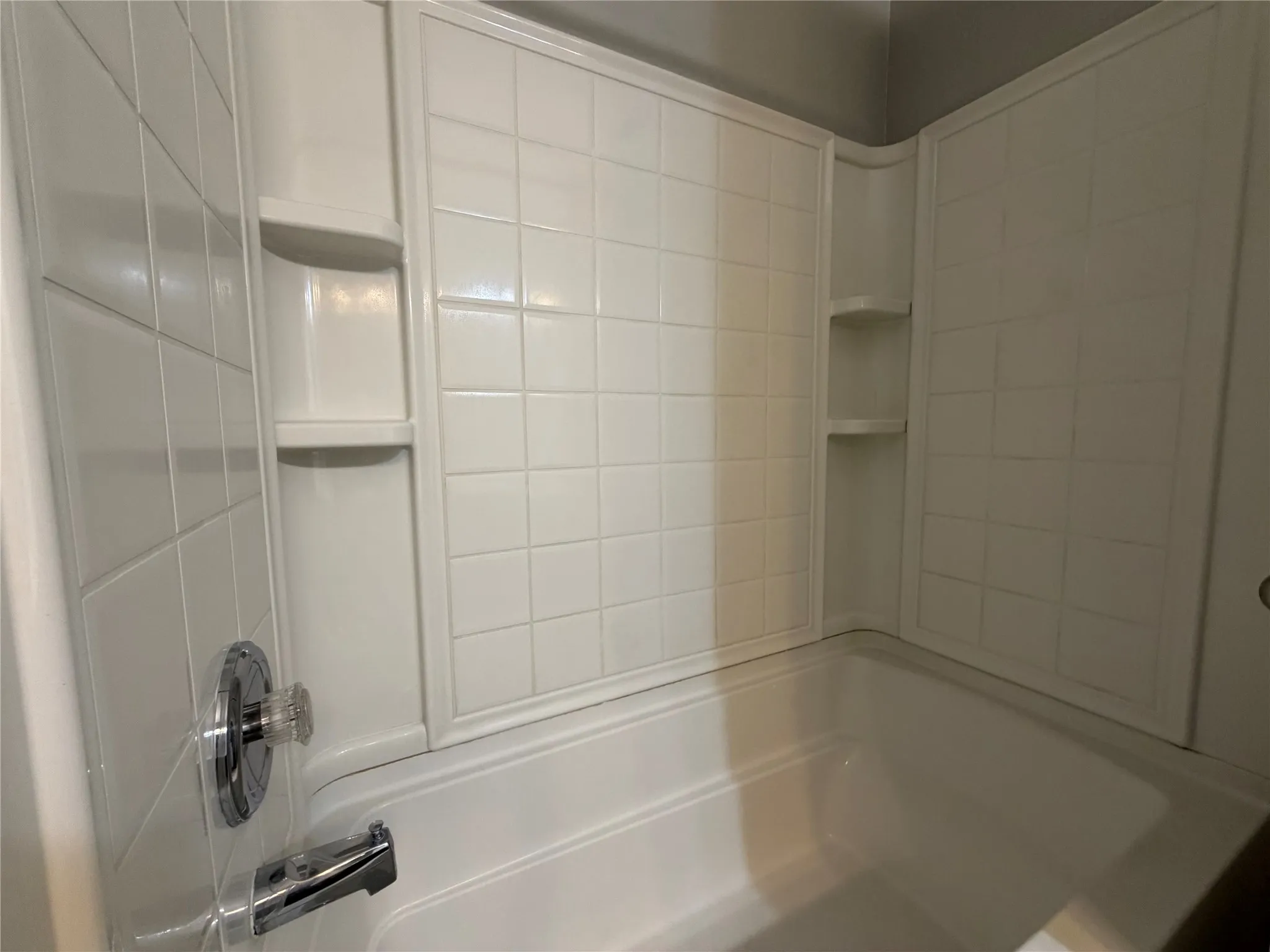 Enclosed bathtub with integrated corner shelving, chrome fixtures, and a white tile-patterned surround
