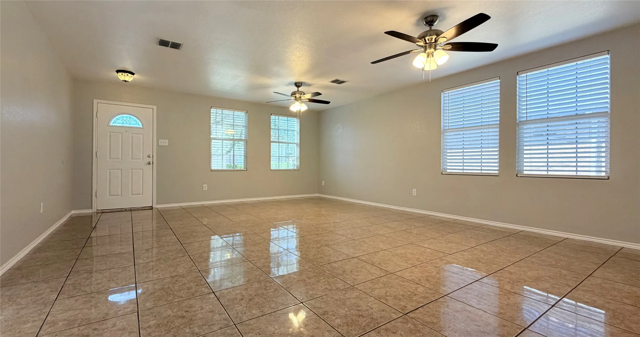 Spacious room featuring tile flooring, multiple windows with blinds, ceiling fans, and a paneled entry door. Living/dining