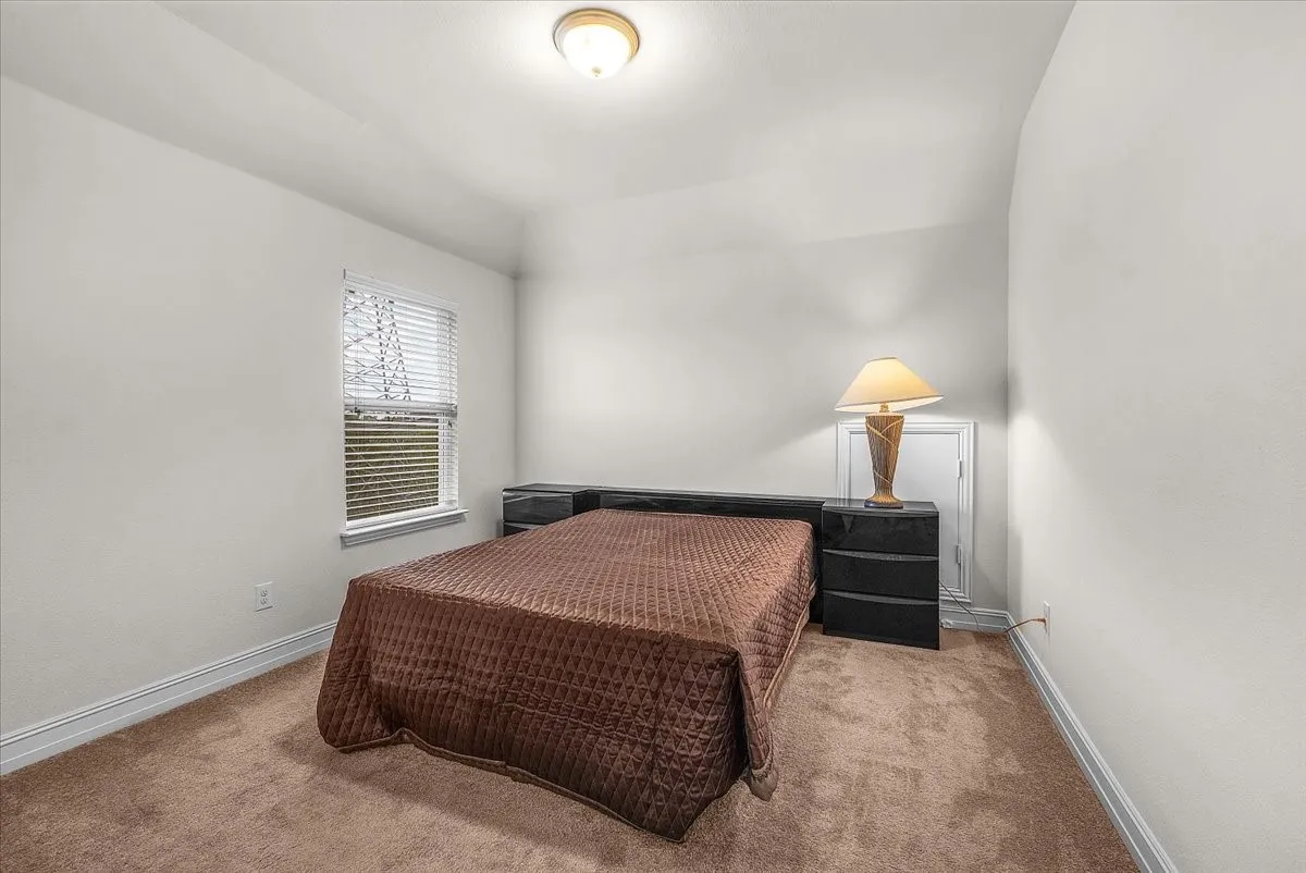 Bright secondary bedroom offering neutral wall finishes, carpeted flooring, a single window with blinds providing natural light, and a ceiling-mounted light fixture