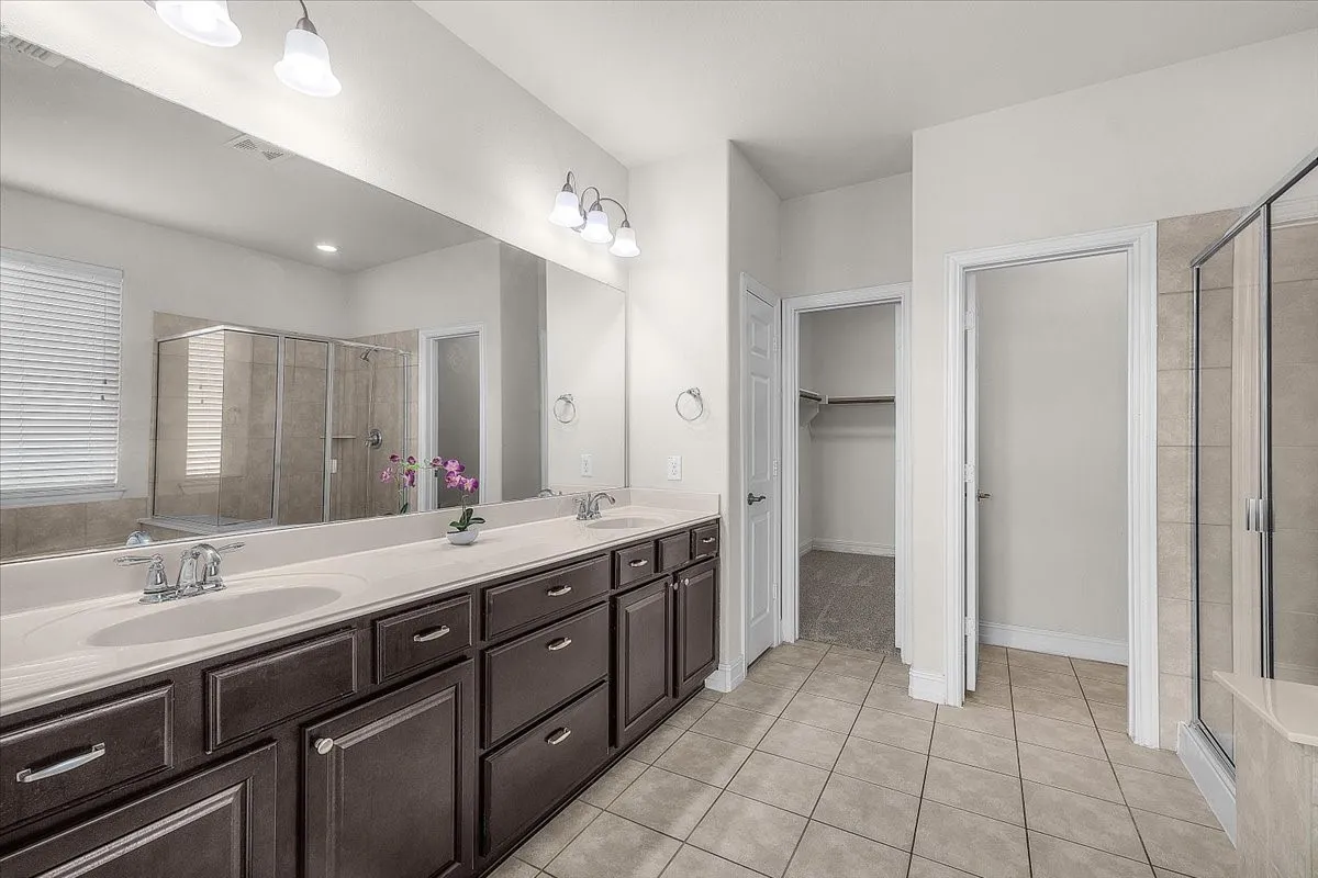 Dual vanity with integrated sinks, expansive mirror, dark wood-finish cabinetry, tile flooring, and a glass-enclosed shower
