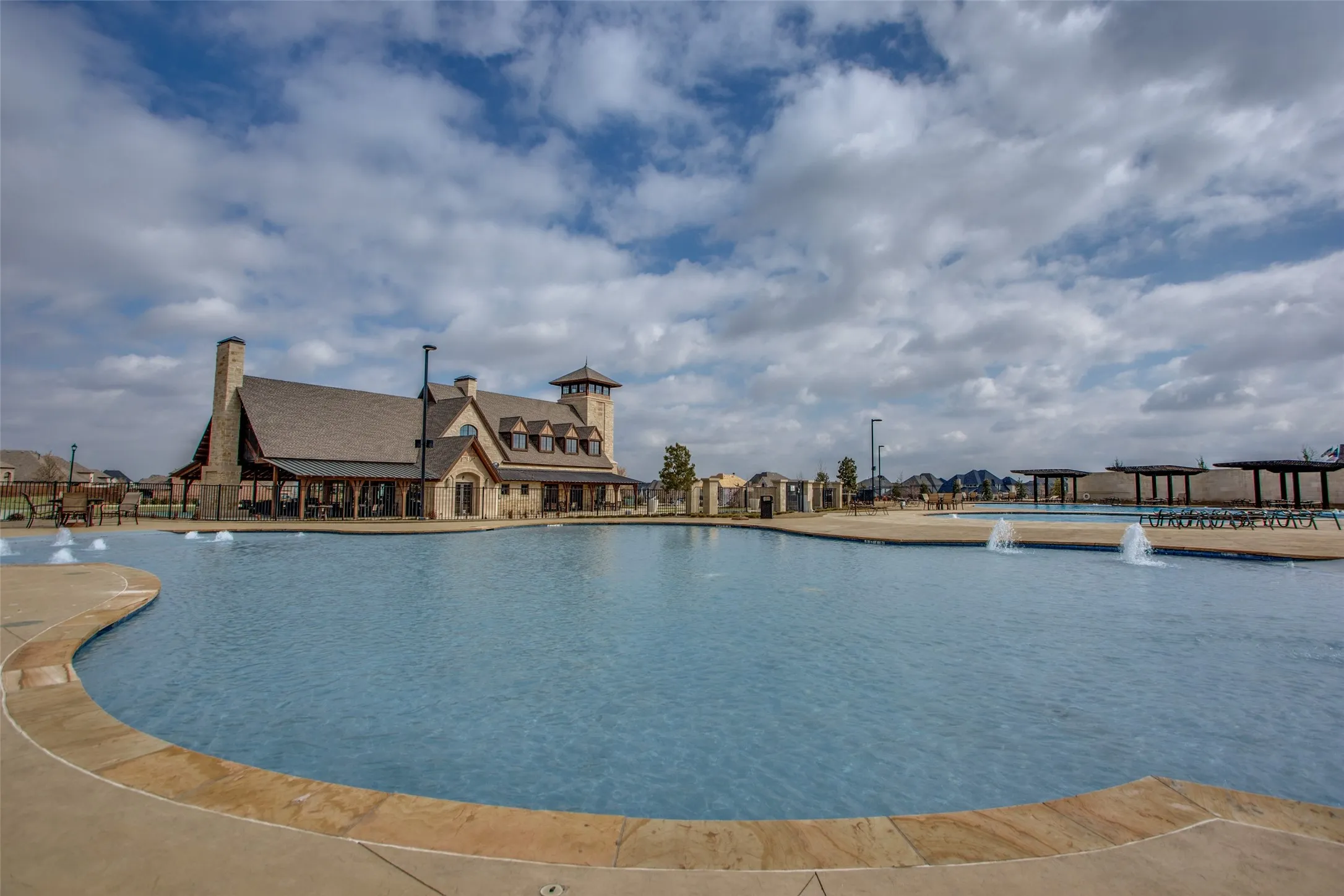 Expansive community pool with integrated fountains, a stone-clad clubhouse featuring a covered patio, and multiple shaded seating areas