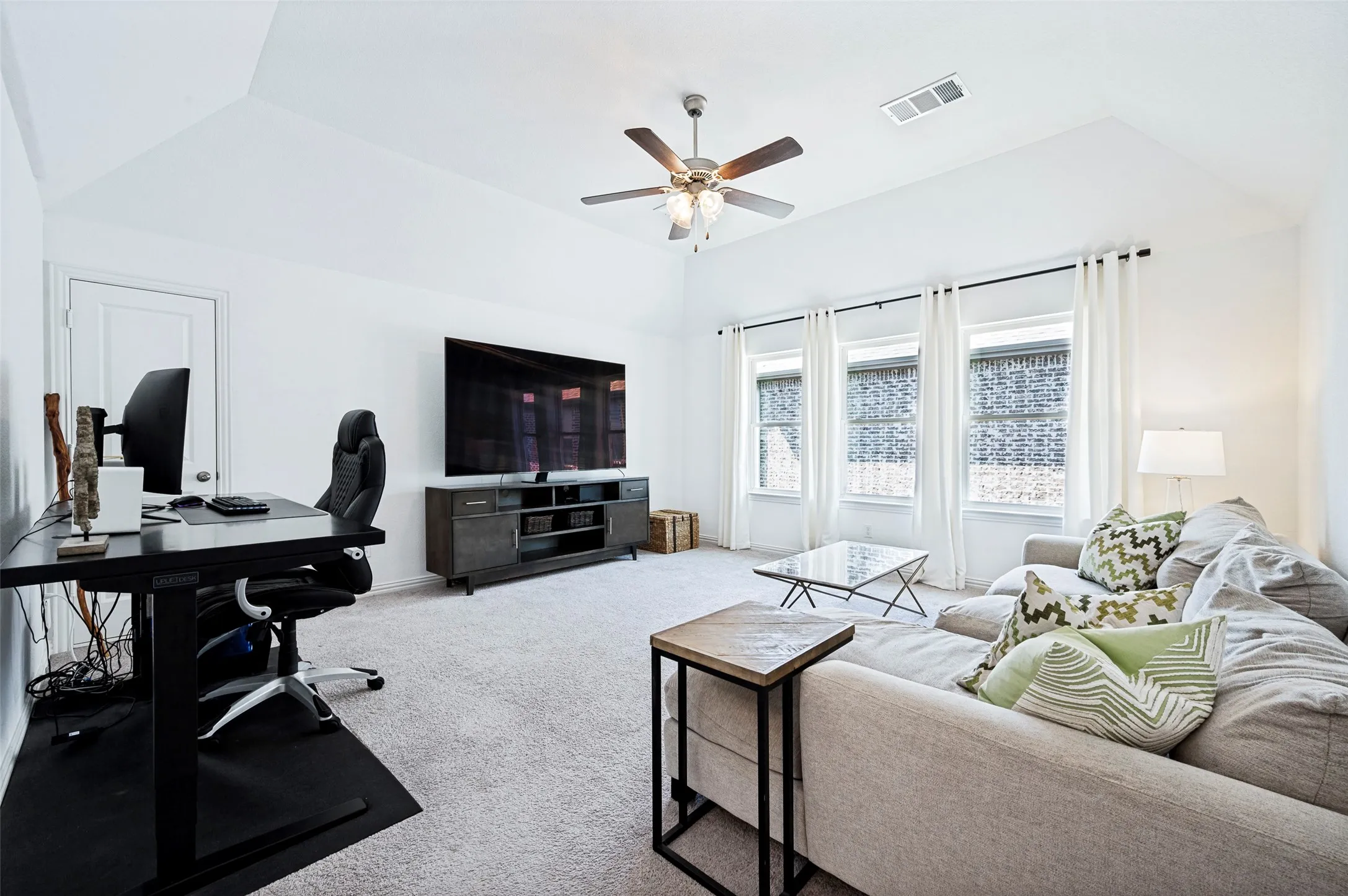 Vaulted ceiling with a ceiling fan, three windows with blinds, light-toned carpeting, and a media console