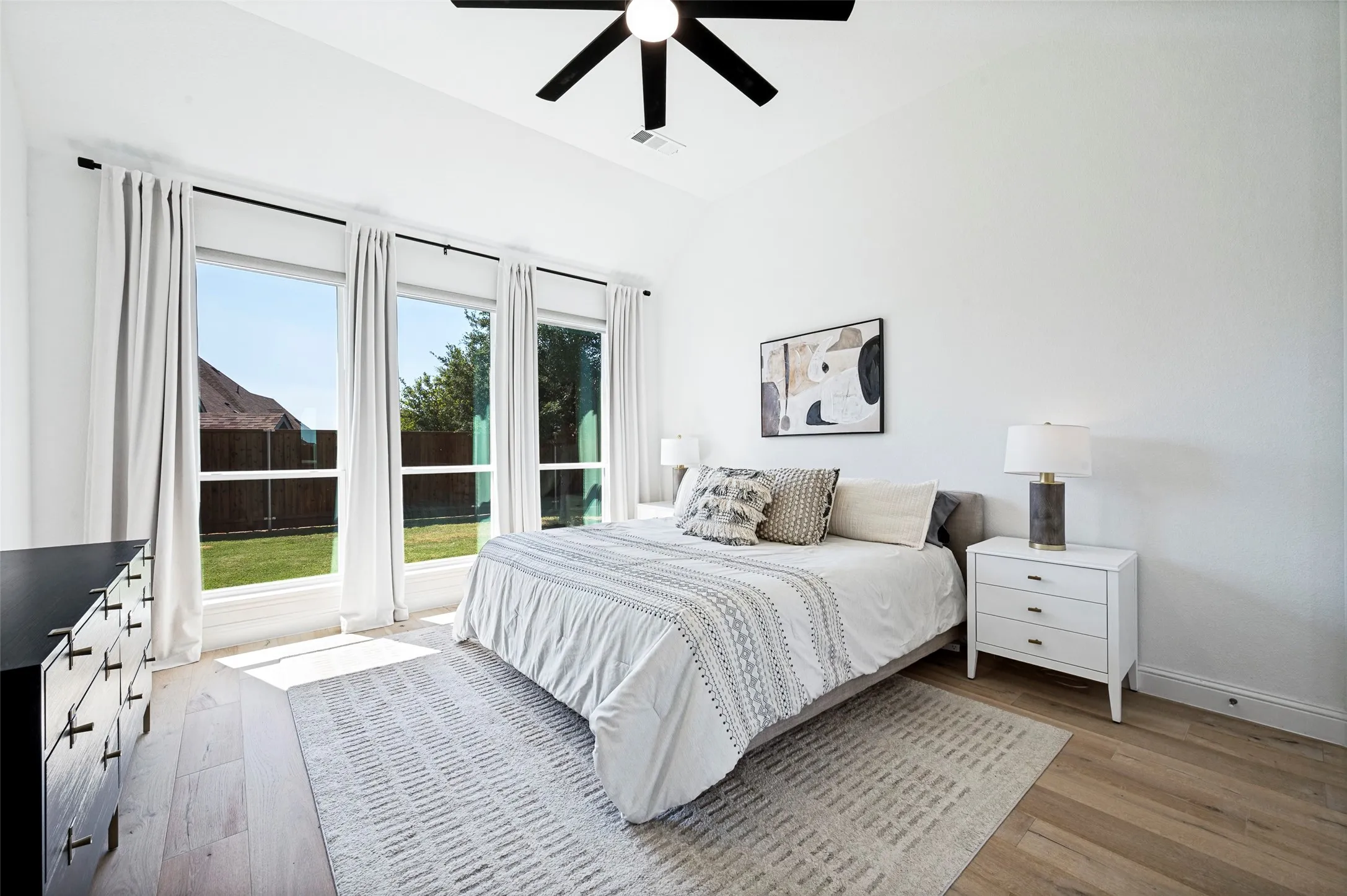 Vaulted ceiling with a black ceiling fan, three large windows, wood-finish flooring, and white walls