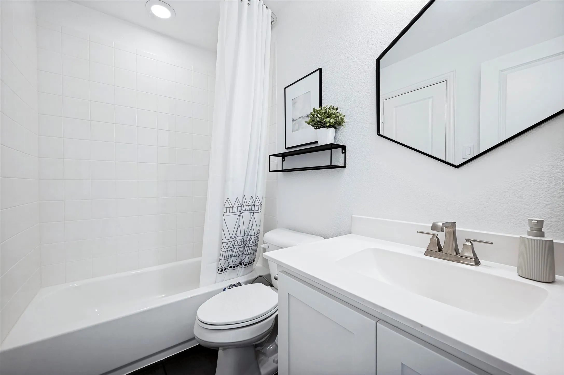 White tile bath surround, hexagon mirror with black frame, white vanity with integrated sink, brushed nickel faucet, and recessed lighting
