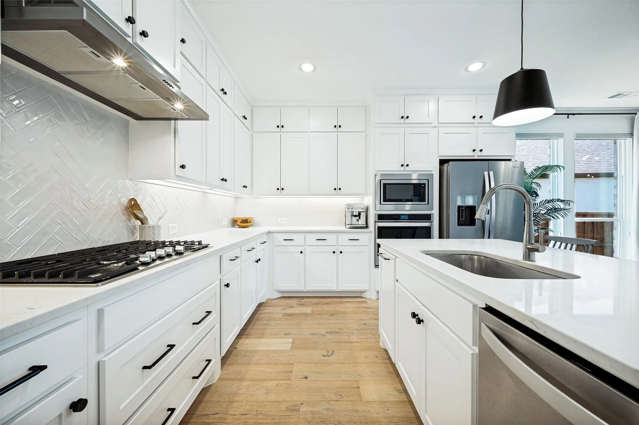 White shaker cabinetry with black hardware, stainless steel appliances, light wood-finish flooring, white countertop surfaces, and a herringbone pattern tile backsplash