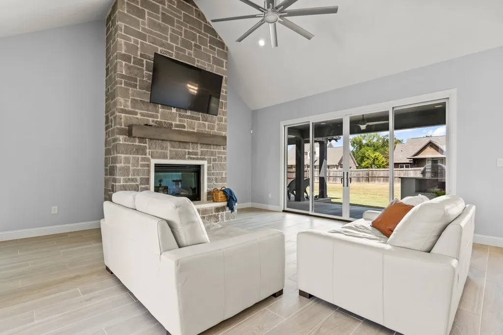 Vaulted ceiling with a ceiling fan, stone fireplace with a wood mantle, large sliding glass doors, and light wood-finish flooring