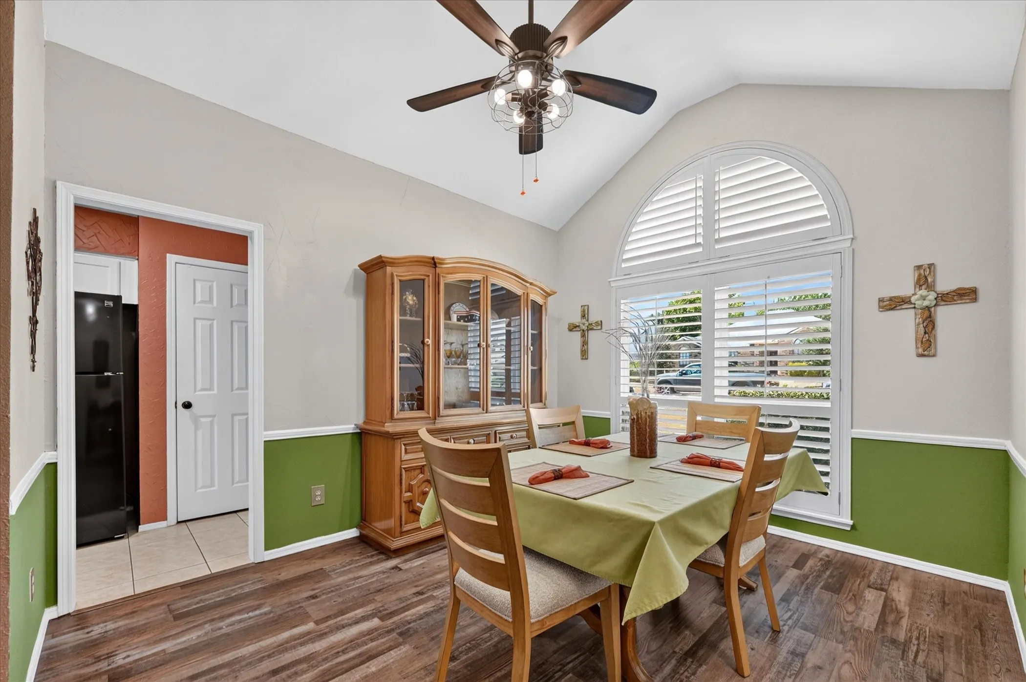 Welcoming dining room features a vaulted ceiling, wood-look flooring, and an impressive arched window with plantation shutters that fills the space with natural light.