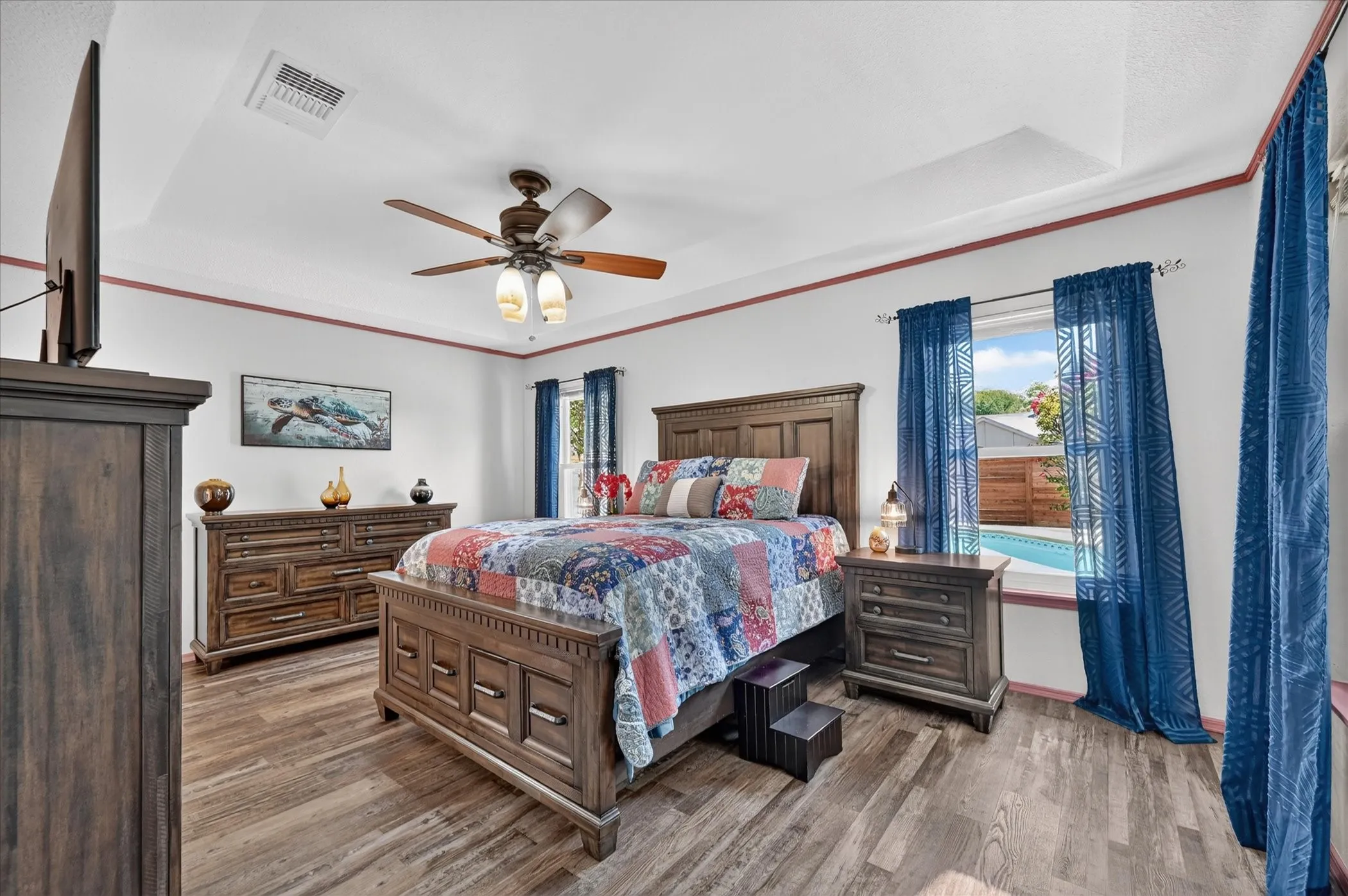 Spacious primary bedroom features a tray ceiling, ceiling fan, wood-look flooring, and multiple windows overlooking the backyard and pool.