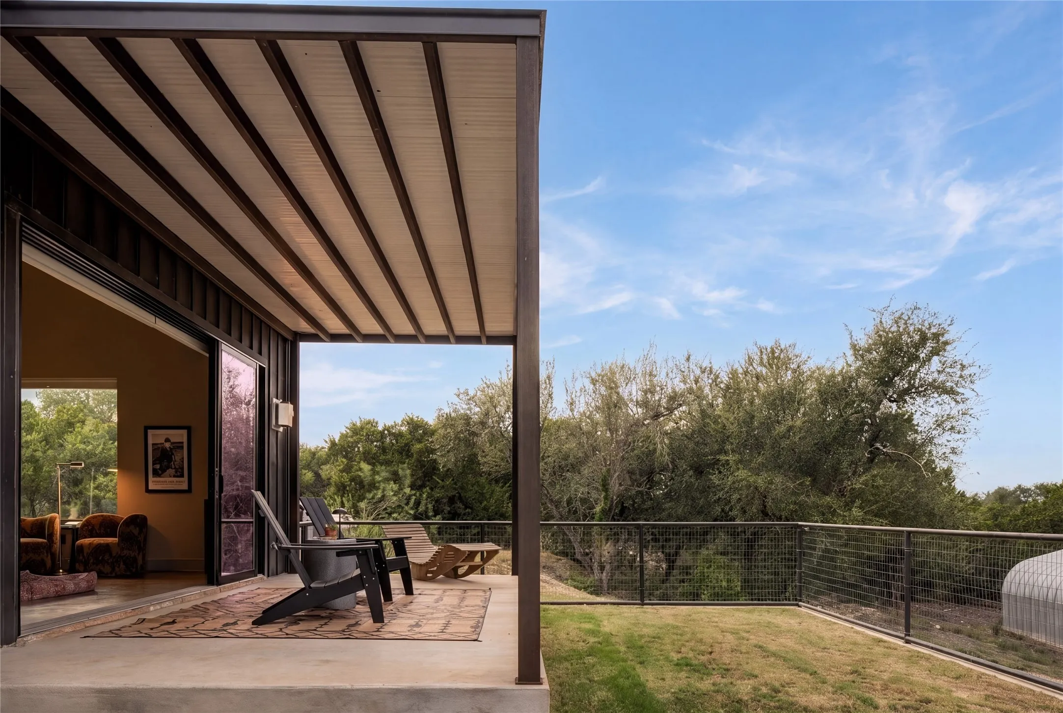 An inviting covered patio borders a grassy yard overlooking lush green trees.
