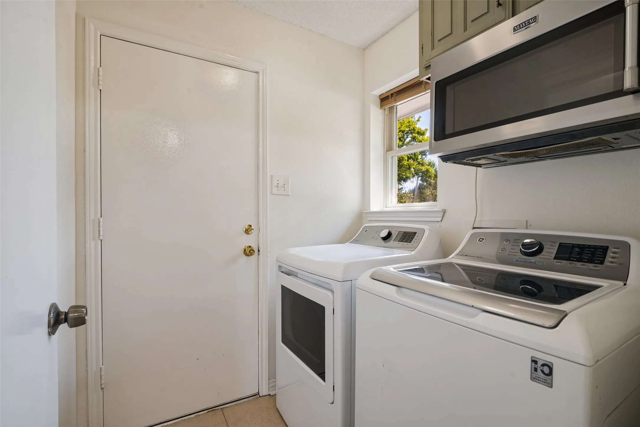Dedicated laundry room featuring side-by-side washer and dryer with stainless steel finishes, ample cabinetry storage above, and a window providing natural light.