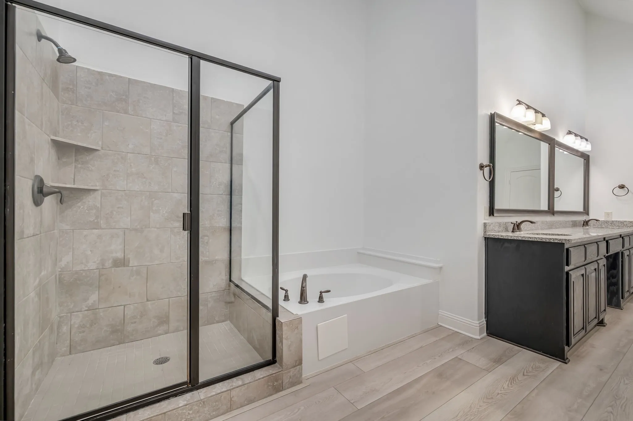 Walk-in shower with tiled walls and built-in shelving, soaking tub, dual vanity with undermount sinks and dark cabinetry, wood-finish flooring, and wall-mounted light fixtures