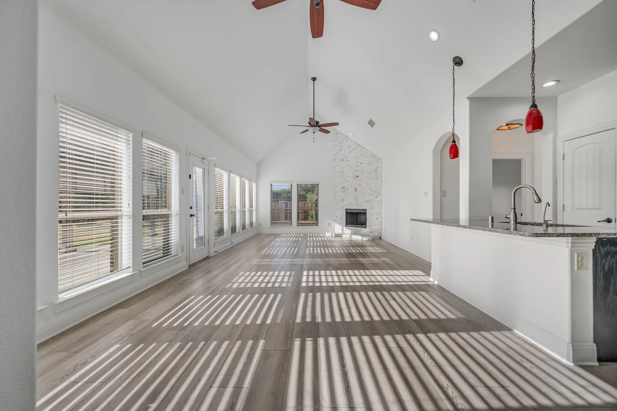 Vaulted ceilings, wood-finish flooring, white brick fireplace, multiple windows with blinds, and a kitchen island with sink and pendant lighting