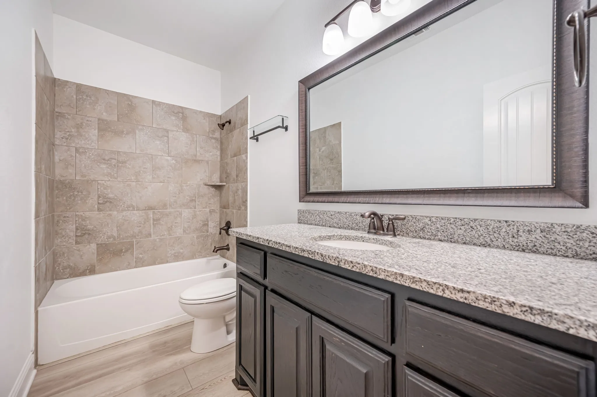 Wood-finish flooring, dark wood-finish vanity with granite countertop, large framed mirror, and a tiled tub and shower combination