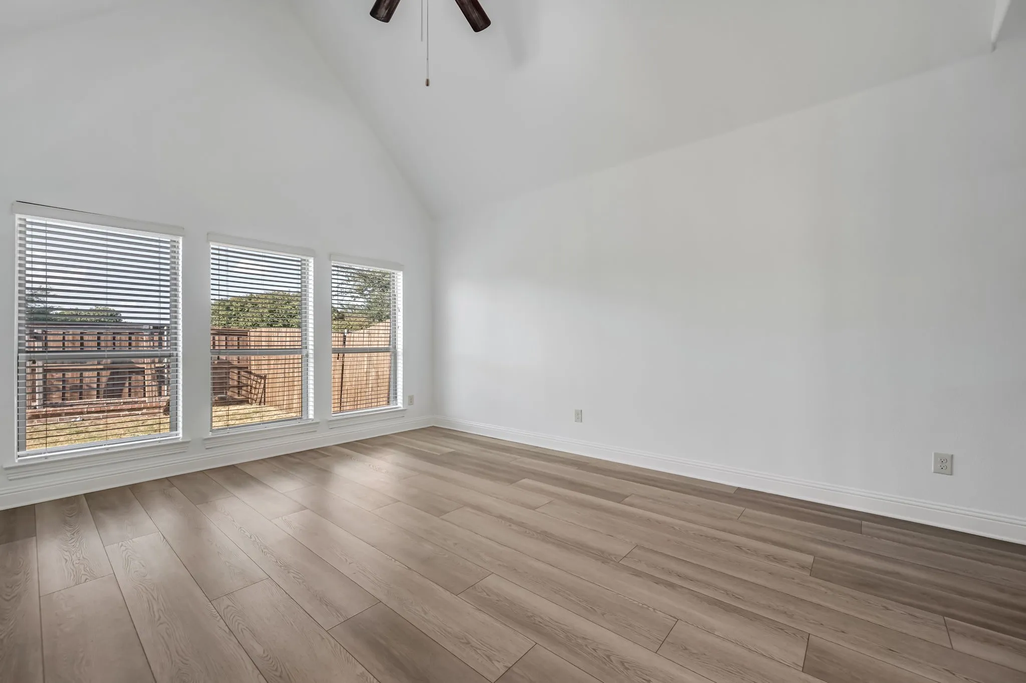 Vaulted ceiling, wood-finish flooring, three large windows with blinds, ceiling fan with light fixture, and white wall finishes