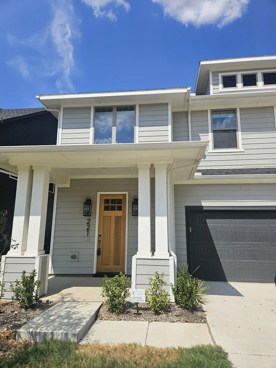 Light gray horizontal siding, prominent white columns, wood-finish entry door, dark garage door, and upper-level windows