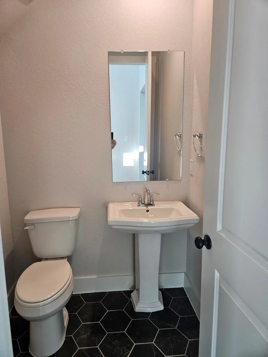 Pedestal sink with chrome faucet, wall-mounted mirror, white toilet, and dark hexagonal floor tiles