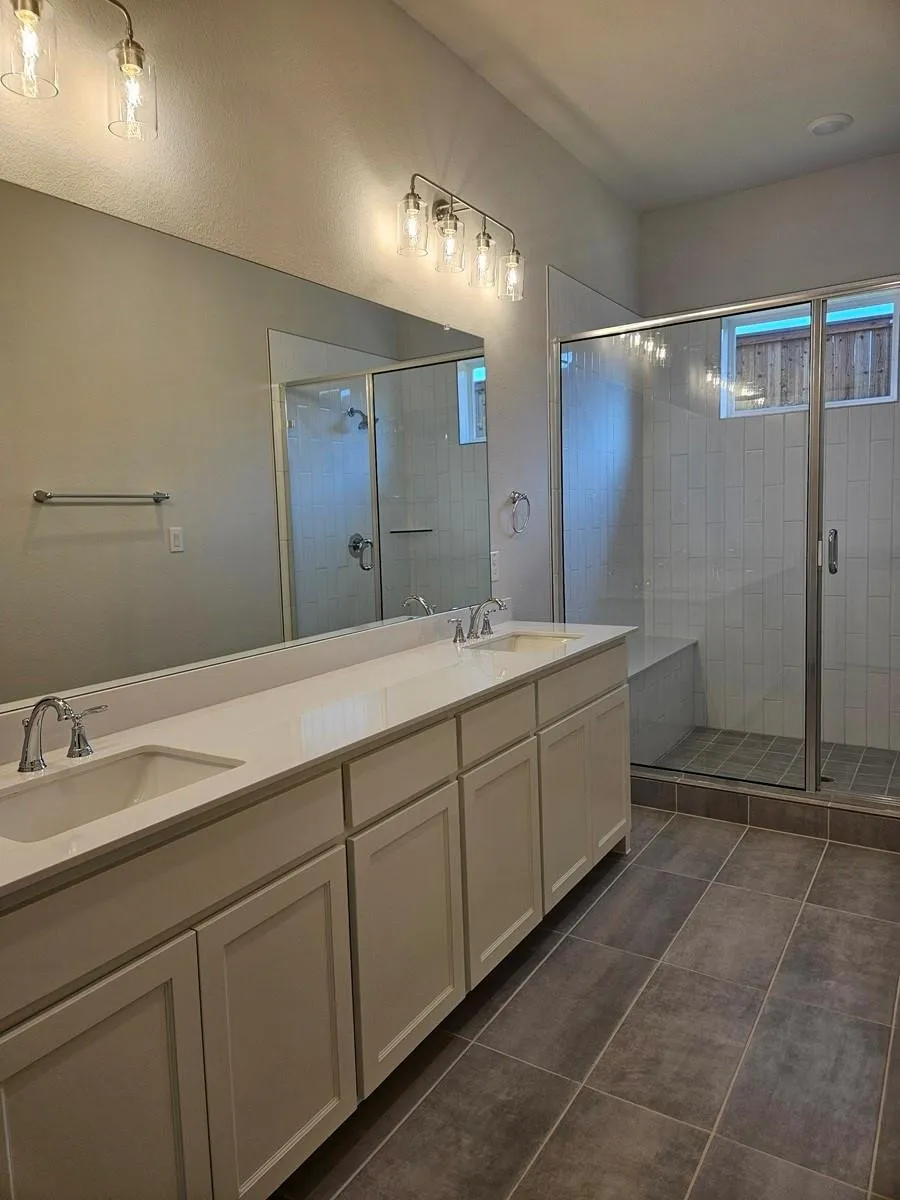Dual vanity with integrated sinks, brushed nickel fixtures, a frameless glass shower enclosure, a built-in shower bench, and large format floor tiling