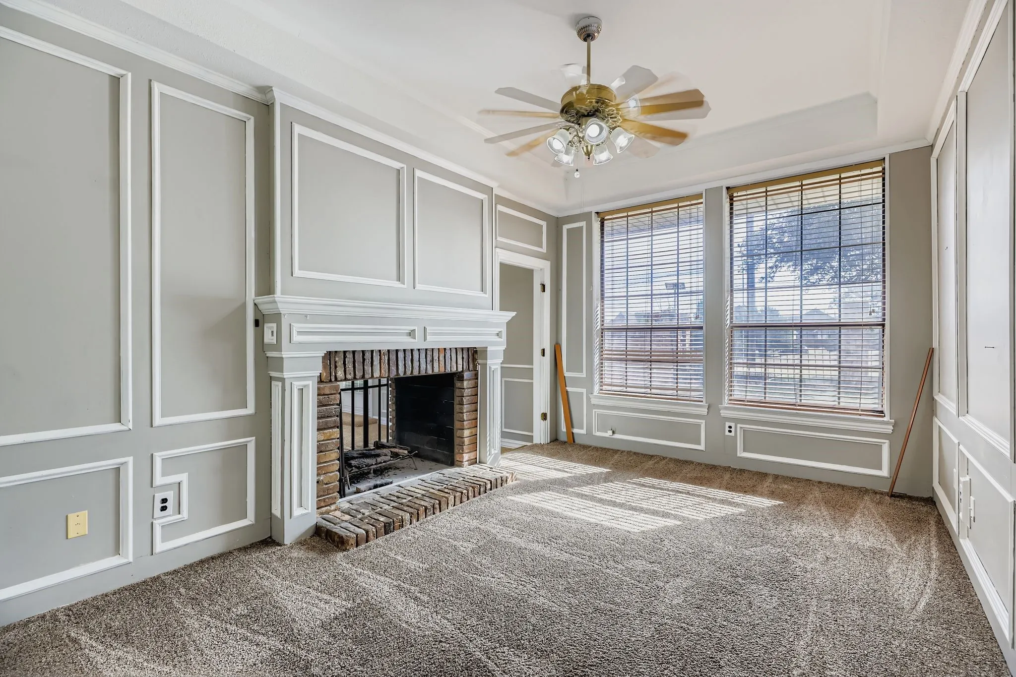 Decorative wall molding, brick fireplace with mantel, tray ceiling, ceiling fan, and two large windows