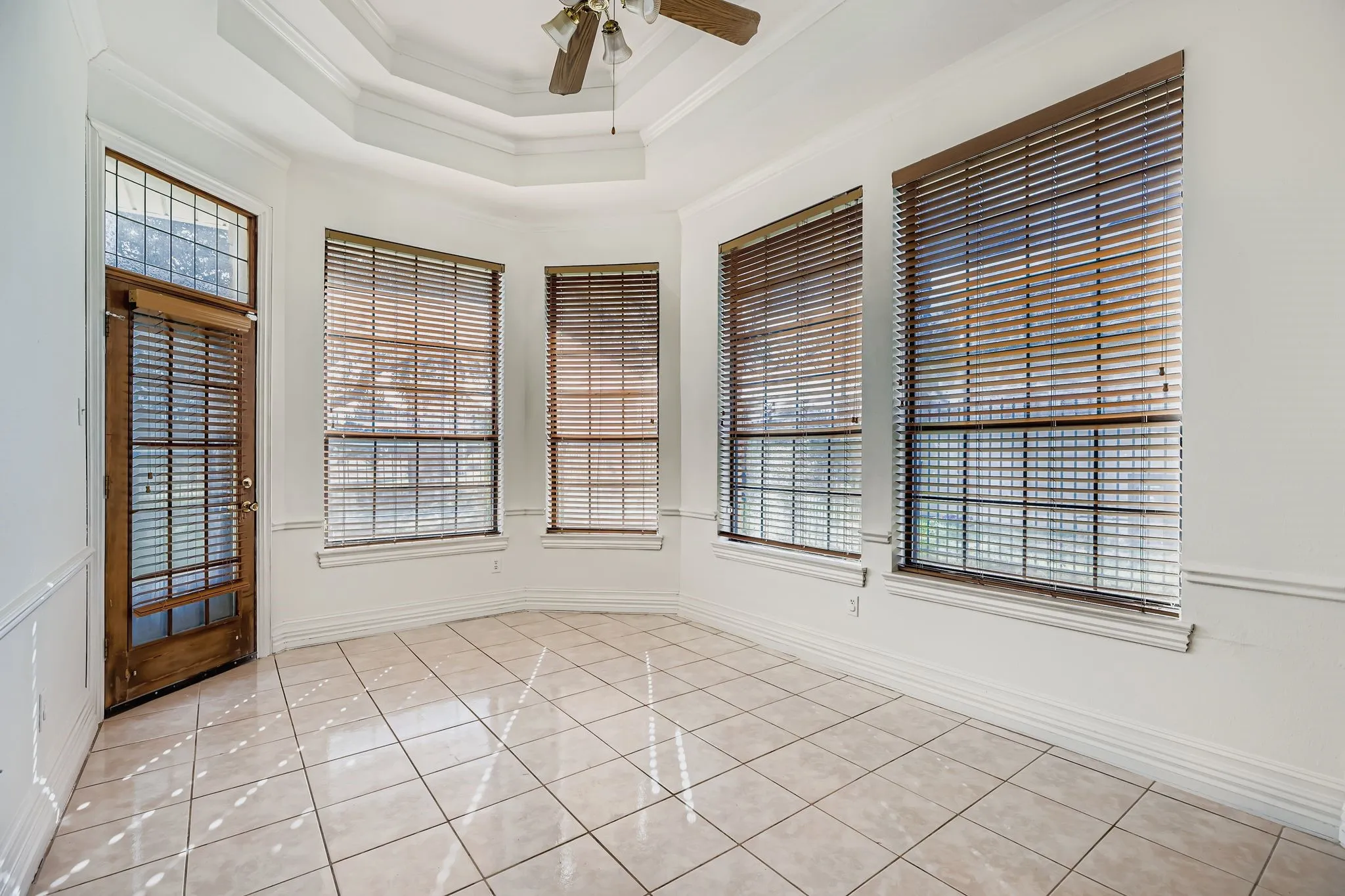 Recessed ceiling with a central fan fixture, multiple large windows with blinds, a wood-paneled door with an upper glass panel, and a tiled floor