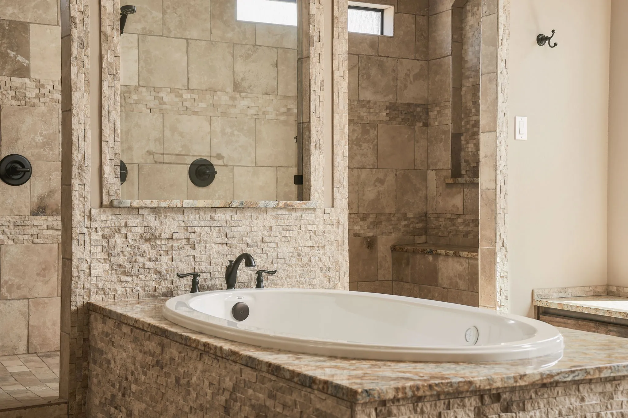 Oval soaking tub with a stone-clad surround, oil-rubbed bronze fixtures, and a tiled walk-in shower featuring a built-in bench