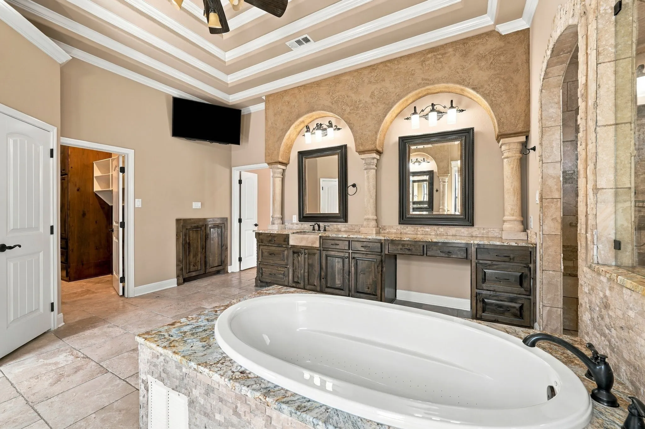Tray ceiling with crown molding, double vanity with dark wood-finish cabinetry, built-in soaking tub with stone tile surround, integrated shelving, and arched architectural details