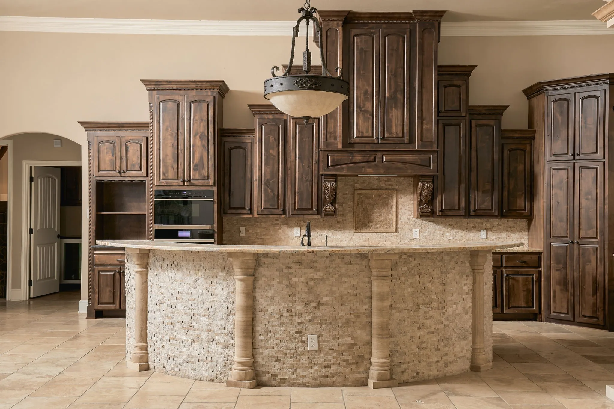 Curved island with stone tile facade, wood-finish cabinetry, integrated wall oven, tile backsplash, and overhead pendant lighting