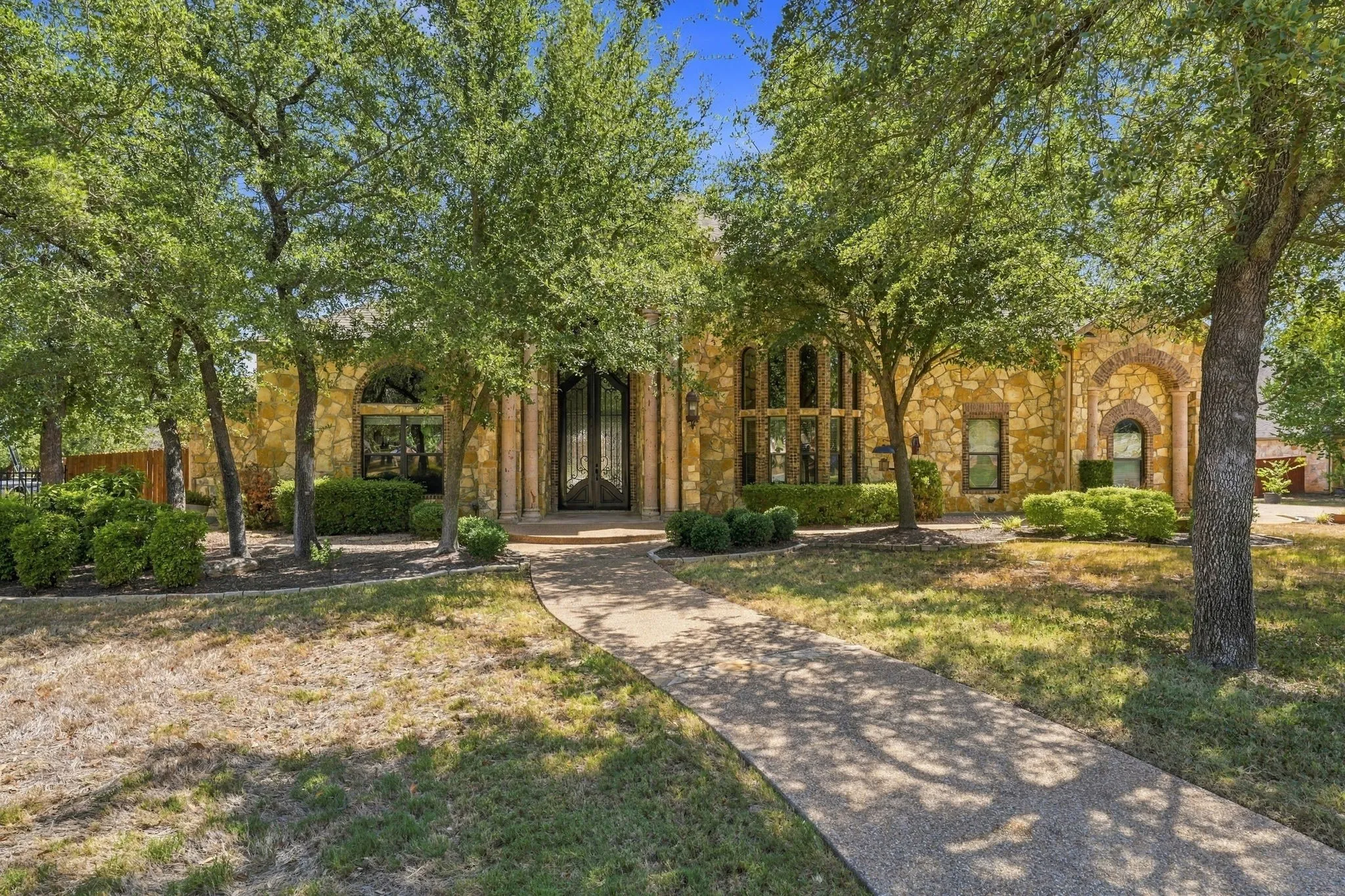 Stone facade exterior with arched windows, wrought iron double entry doors, mature trees, and a concrete pathway