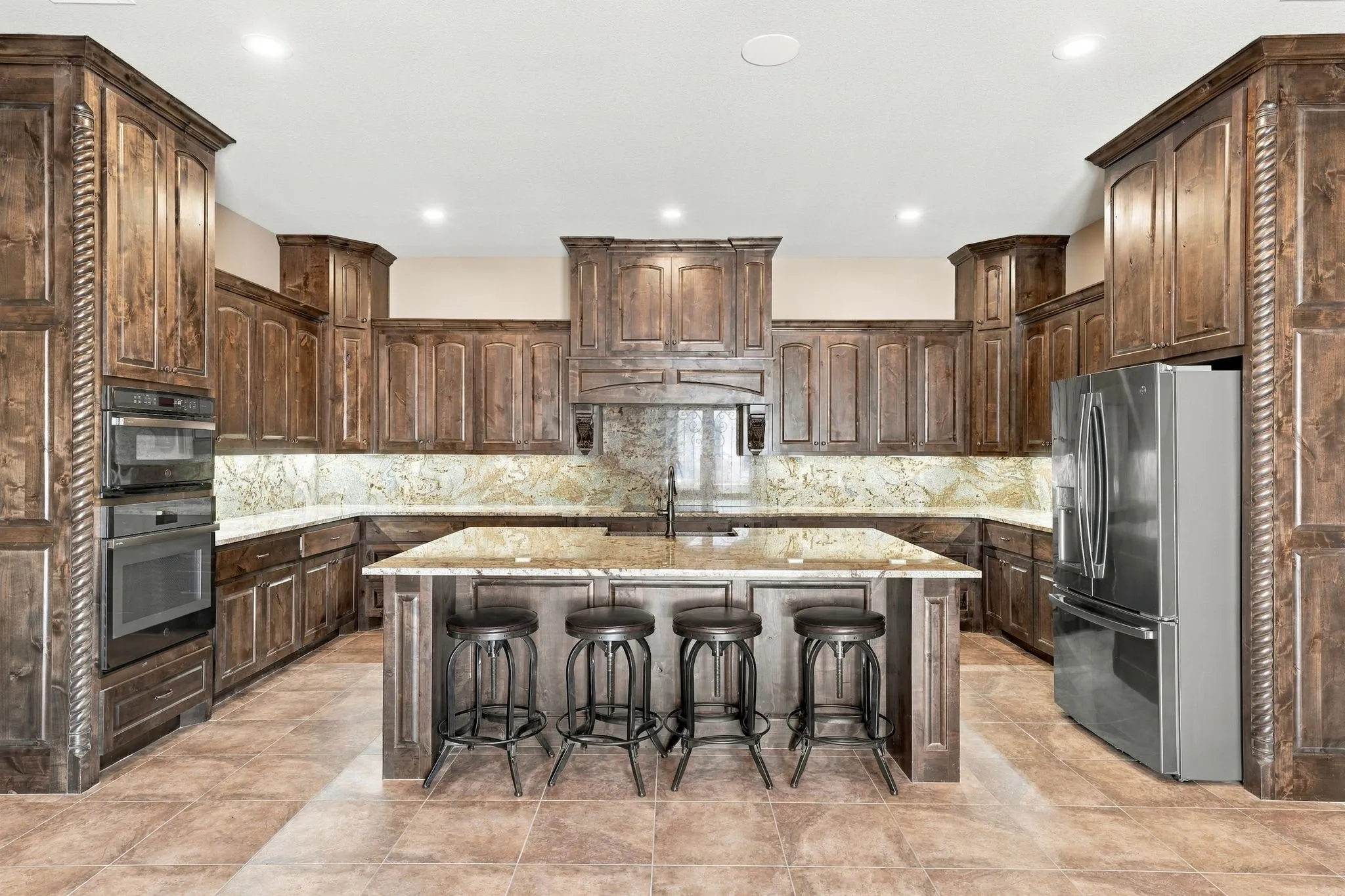Gourmet kitchen with rich wood cabinetry, built-in double ovens, a large central island, stone countertops, and a tiled backsplash