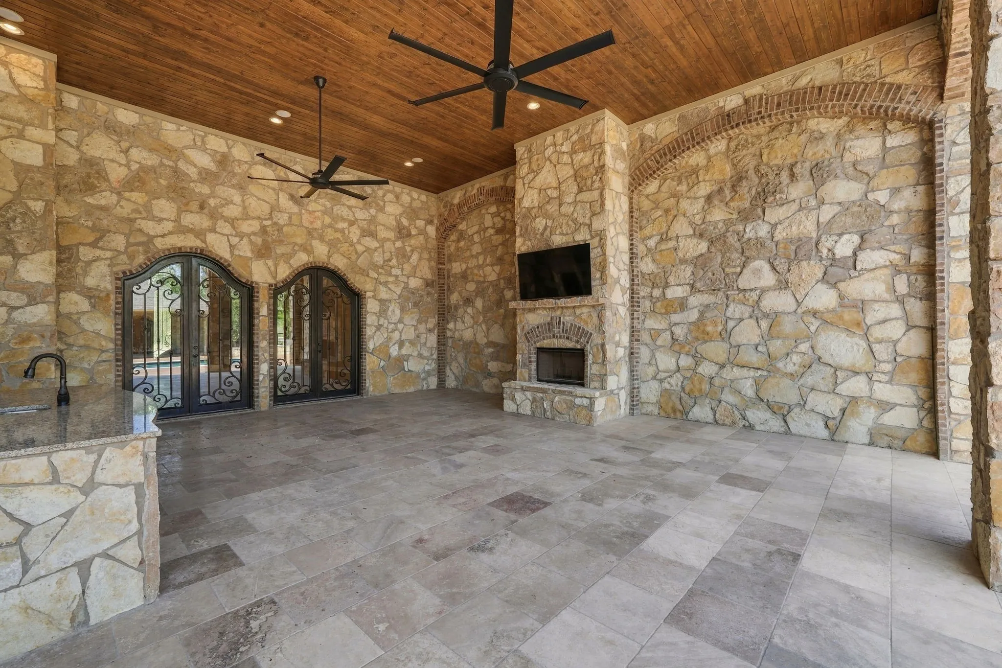 Expansive covered patio featuring stone walls, a wood-paneled ceiling, large format tile flooring, a stone fireplace, and built-in outdoor kitchen with a sink