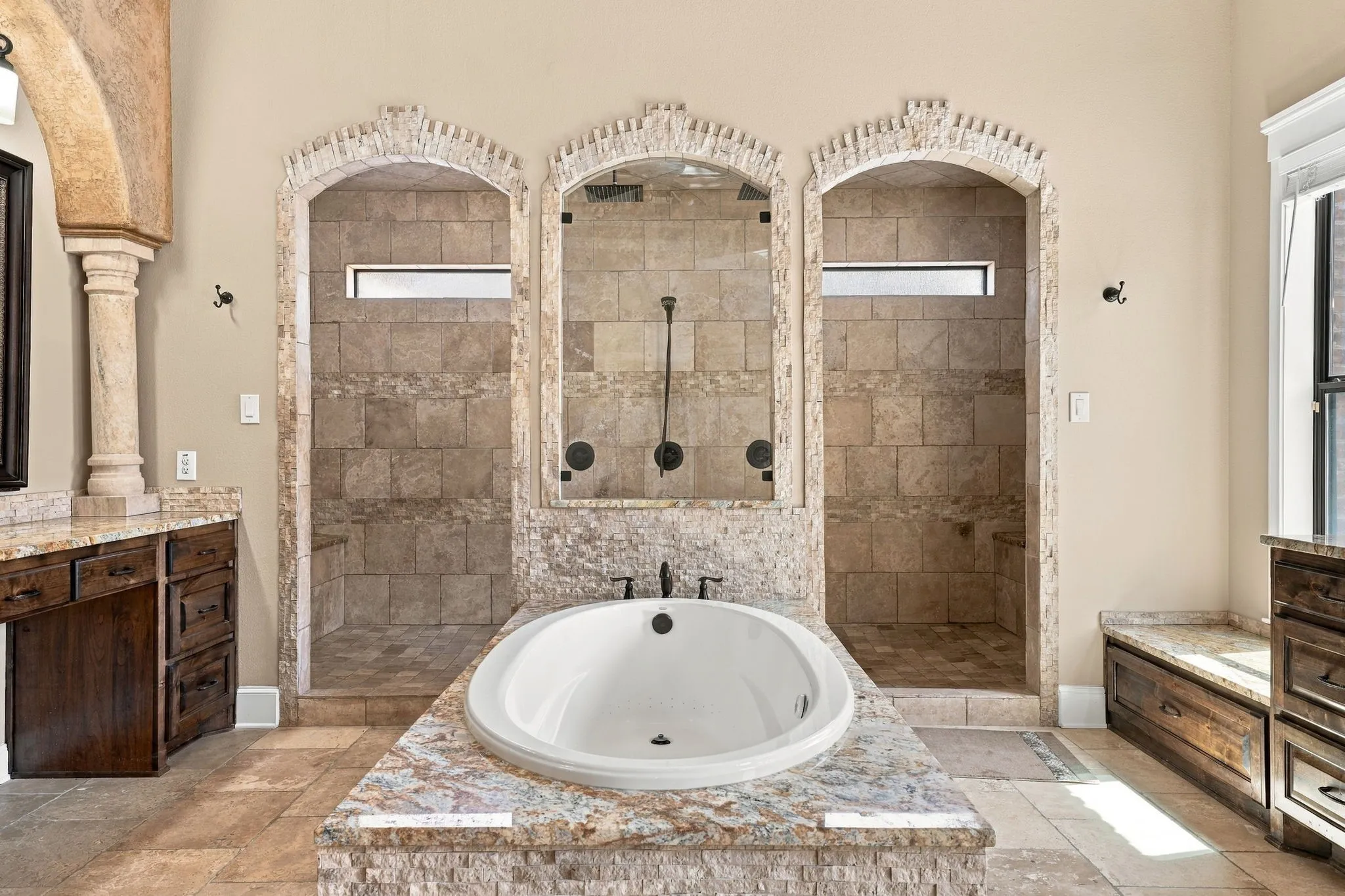 Oval soaking tub set in a stone surround, dual stone-tiled showers with arched entryways, stone-tiled flooring, and a window with natural light