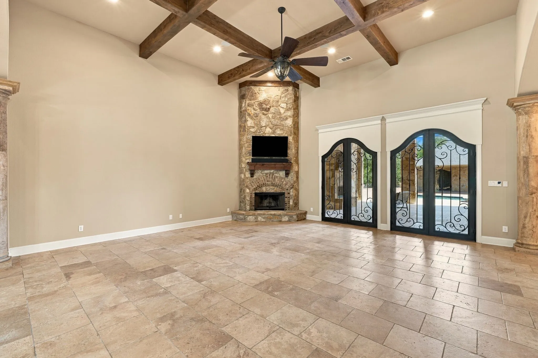 Vaulted ceilings with exposed wood beams, stone fireplace, ceiling fan with light fixture, tile flooring, and custom arched glass doors with wrought iron detailing