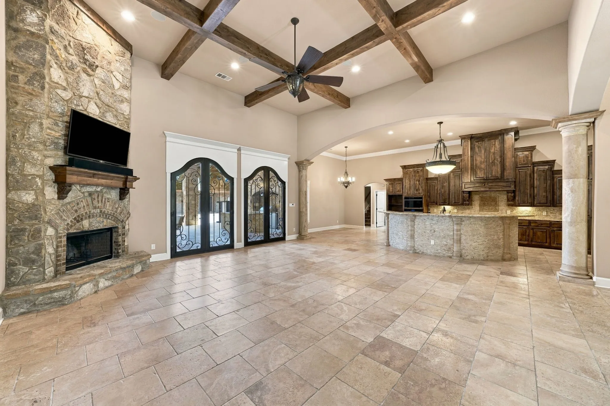 Vaulted ceiling with exposed wood beams, stone fireplace, wood-finish cabinetry, tile flooring, and decorative wrought iron doors