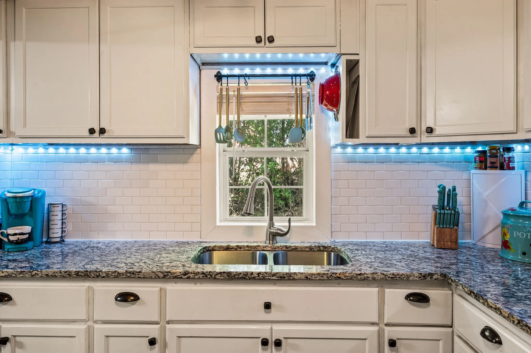 White shaker cabinetry with dark hardware, undermount double basin sink with gooseneck faucet, light granite countertops, white subway tile backsplash, and task lighting