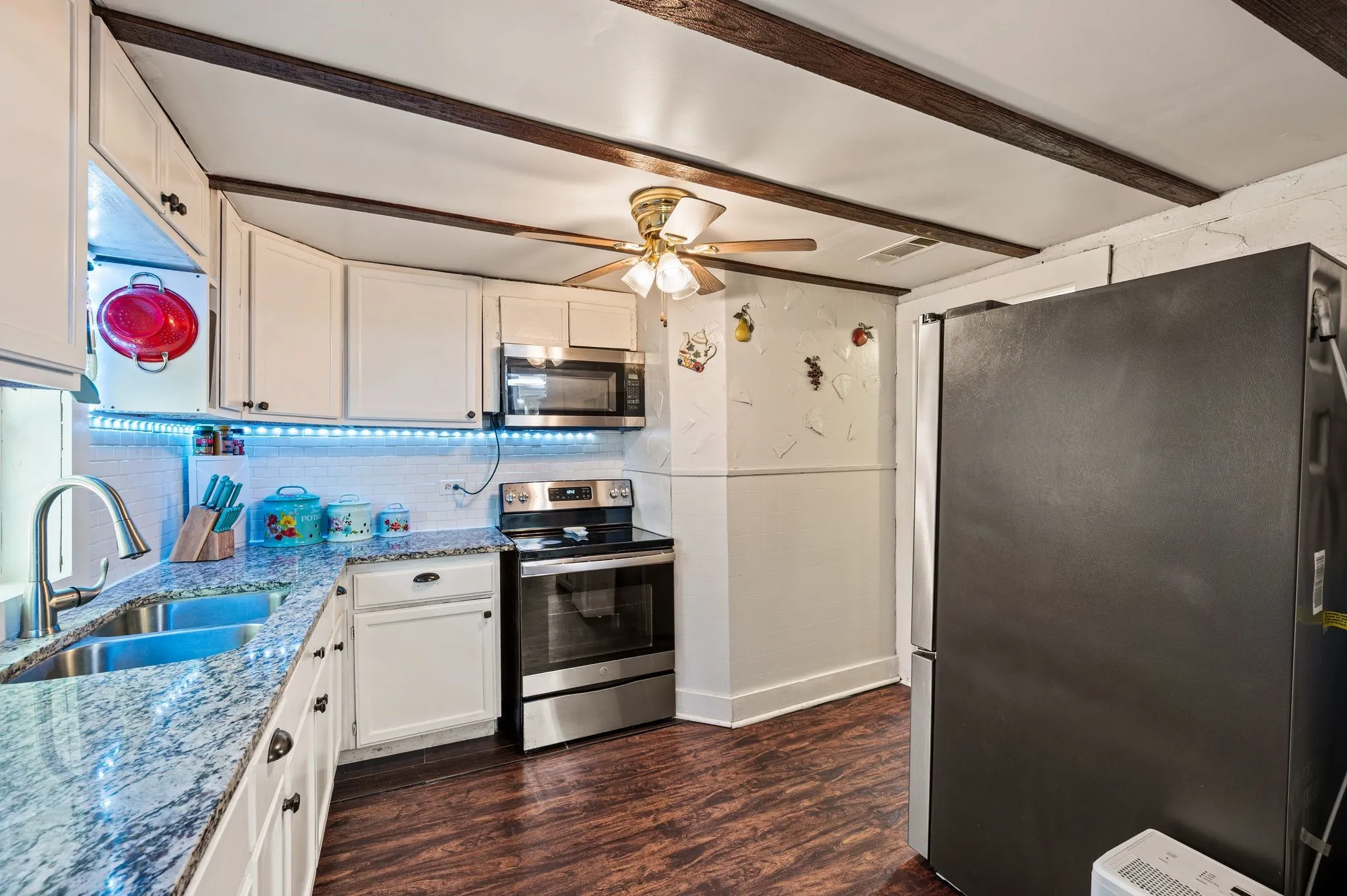 Kitchen featuring white cabinetry, wood-finish flooring, a double basin stainless steel sink, stainless steel appliances, and exposed ceiling beams
