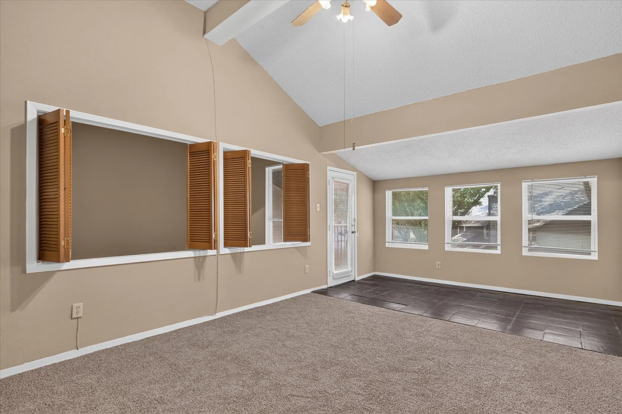 Vaulted ceiling with ceiling fan, large interior window with wood shutters, multiple exterior windows, tile flooring, and carpet flooring
