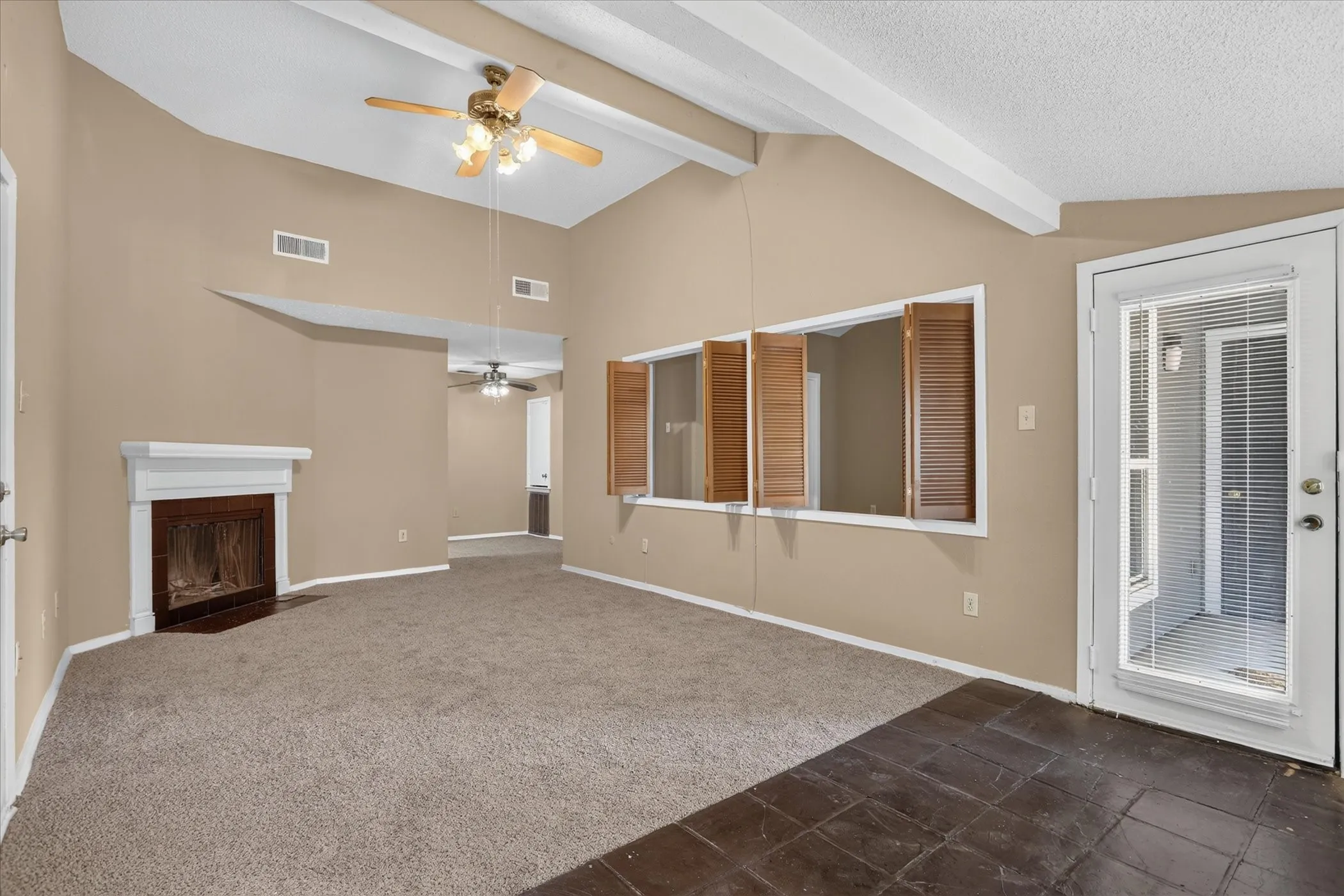 Vaulted ceiling with exposed beams, ceiling fan, fireplace with white mantel, windows with wooden shutters, and a glass-paneled exterior door