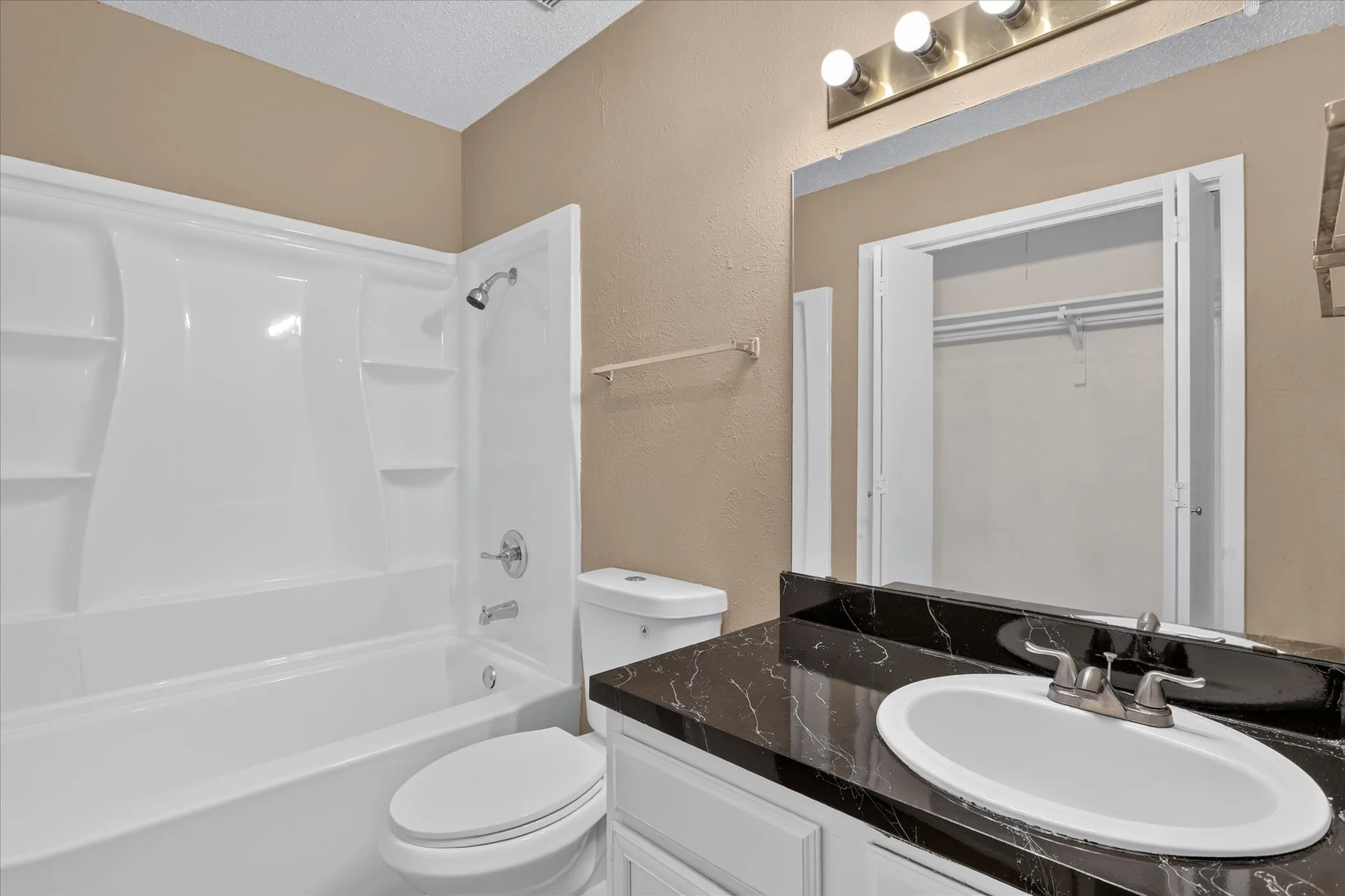 White bathtub with integrated shower, single vanity with oval sink, dark countertop with light veining, and wall-mounted mirror
