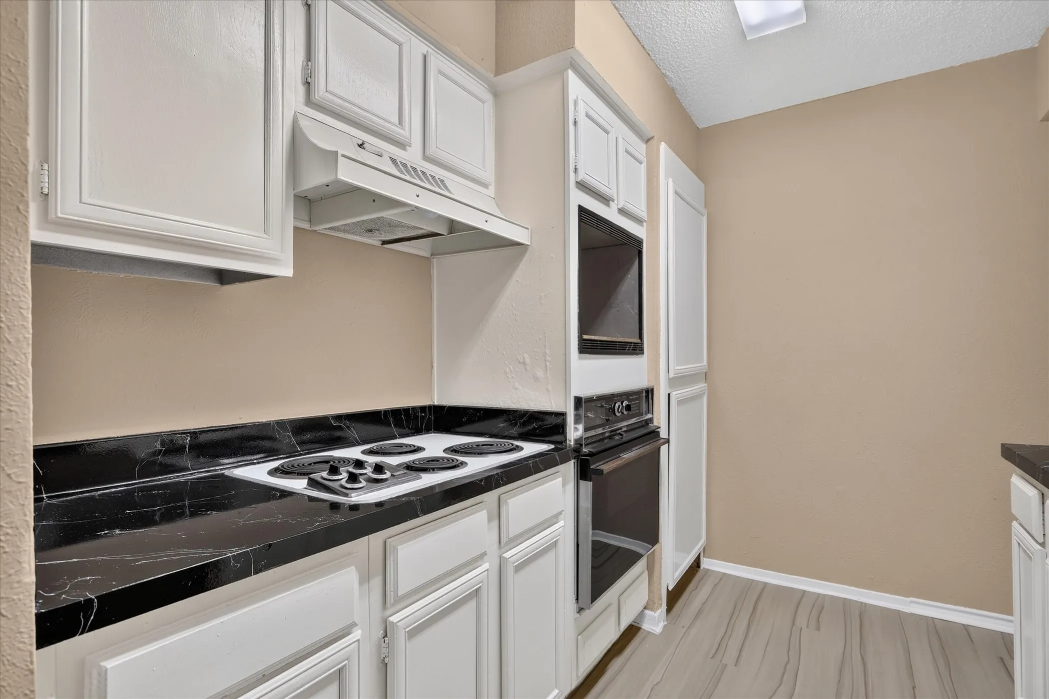 White cabinetry, black countertops, built-in oven, built-in microwave, and wood-finish flooring