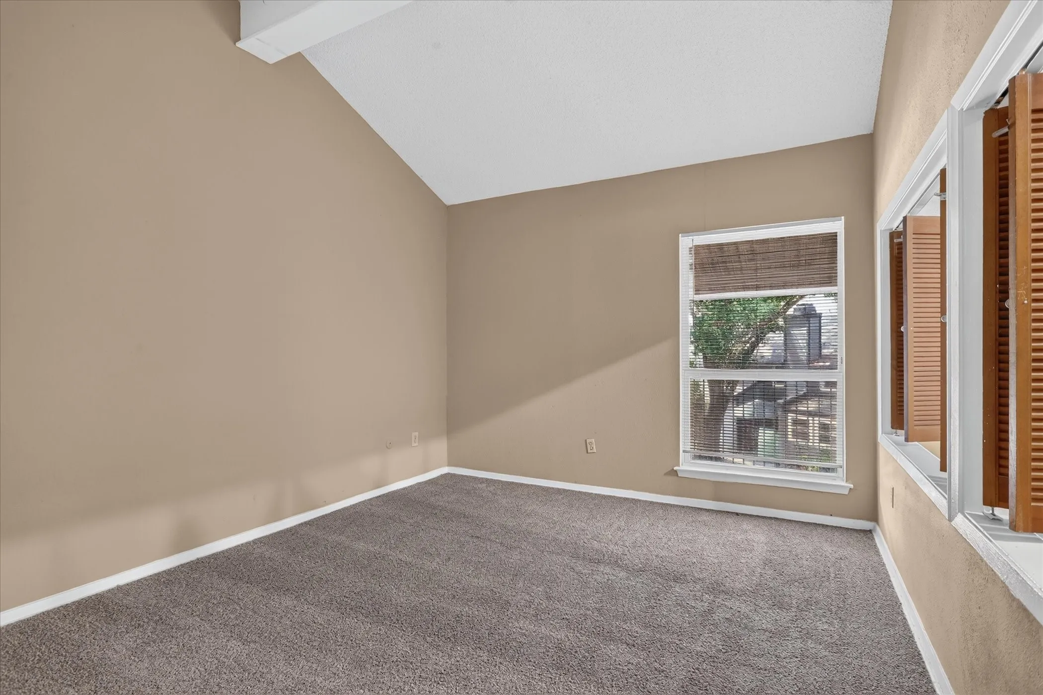 Vaulted ceiling, neutral wall tones, gray carpet, double-hung window, and wooden louvered shutters