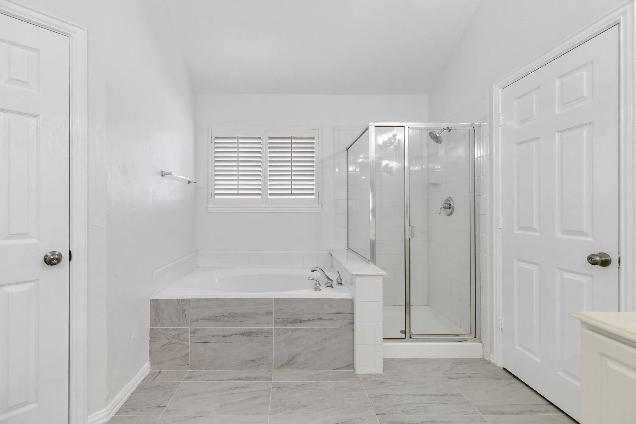 Built-in soaking tub with tile surround, glass-enclosed shower, louvered window, and light-toned tile flooring