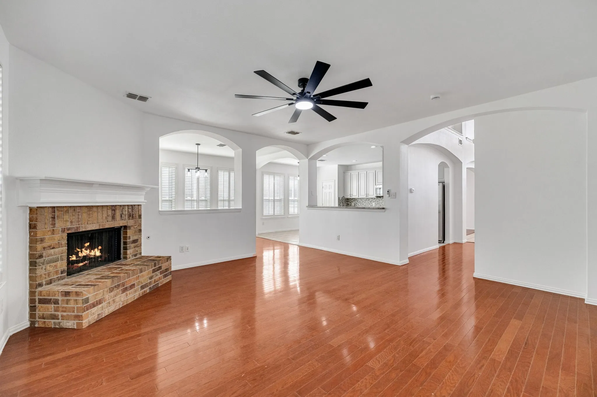 Wood-finish flooring, brick fireplace with mantel, arched doorways, white cabinetry, and a ceiling fan with integrated lighting